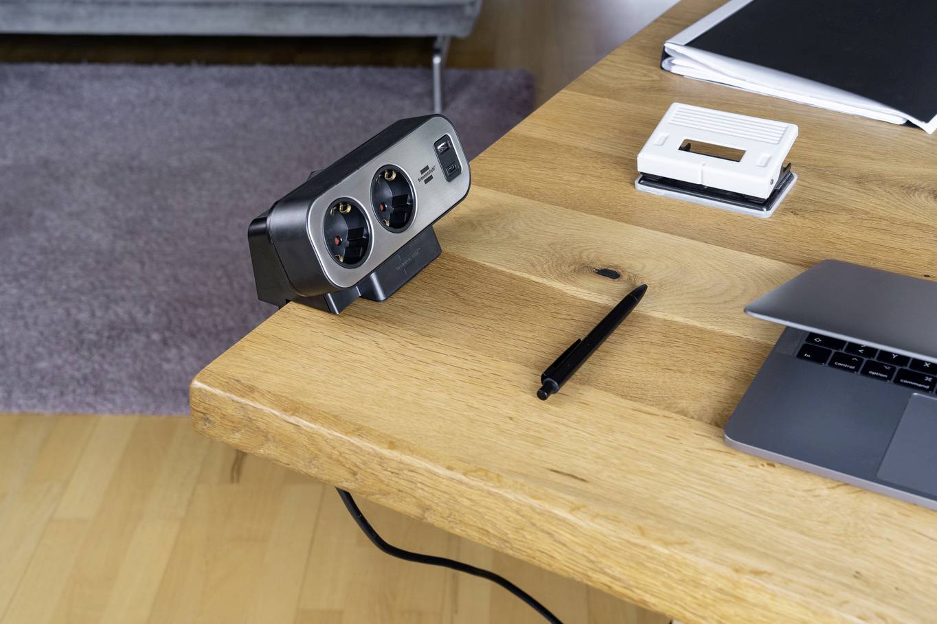 Auf einem Holztisch liegen ein Laptop, ein Kugelschreiber, und eine Steckdosenleiste. Im Hintergrund ist ein Teppich zu sehen.