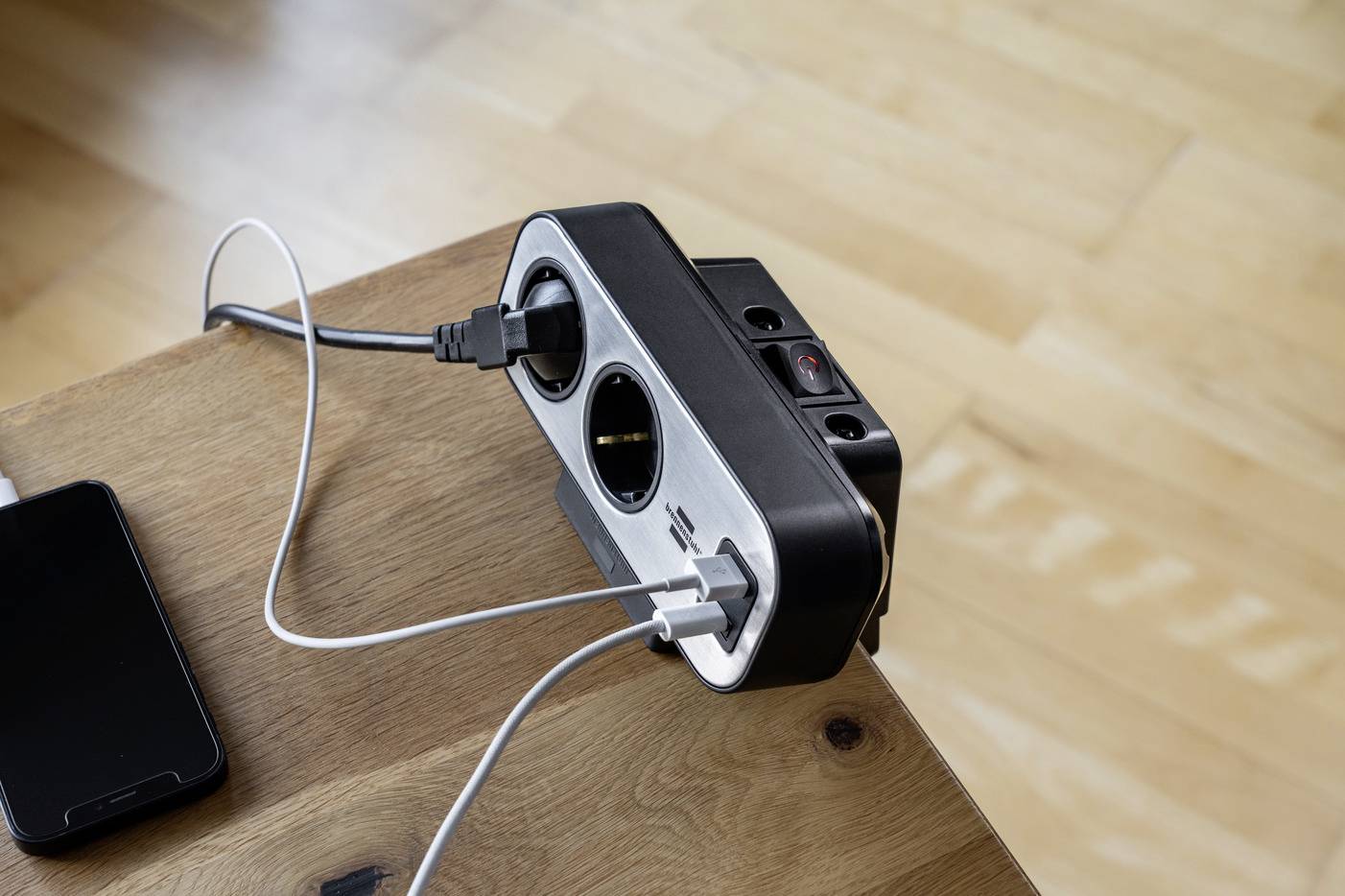 Mehrfachsteckdose auf Holztisch, angeschlossen sind ein Mobiltelefonladegerät und ein anderes elektrisches Gerät, Holzboden im Hintergrund.