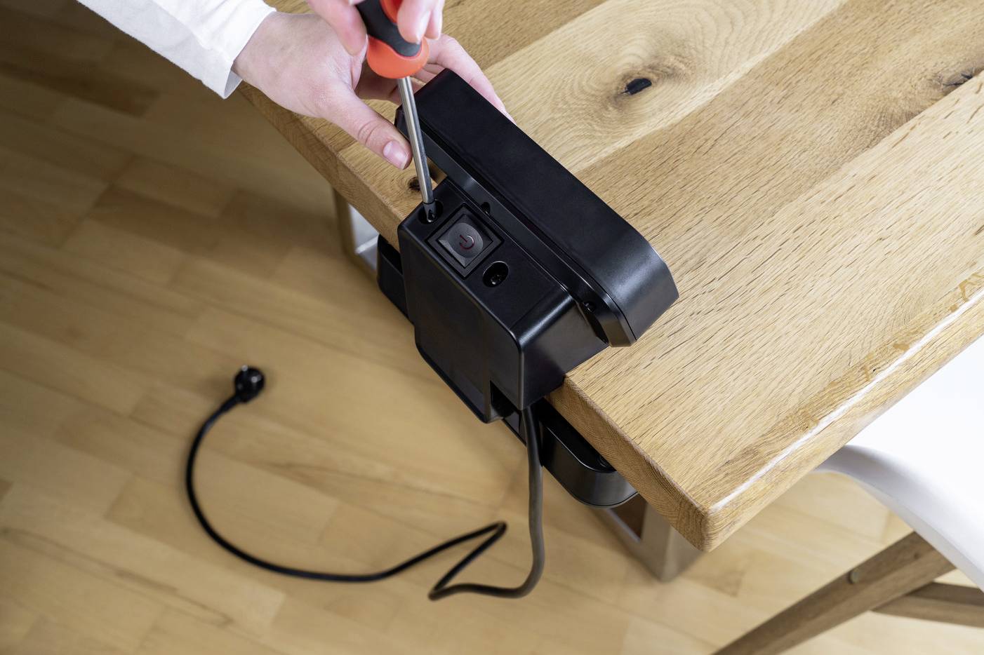 Eine Person montiert eine schwarze Steckdosenleiste an einem Holztisch mit einem Schraubendreher. Ein Stromkabel hängt herab.