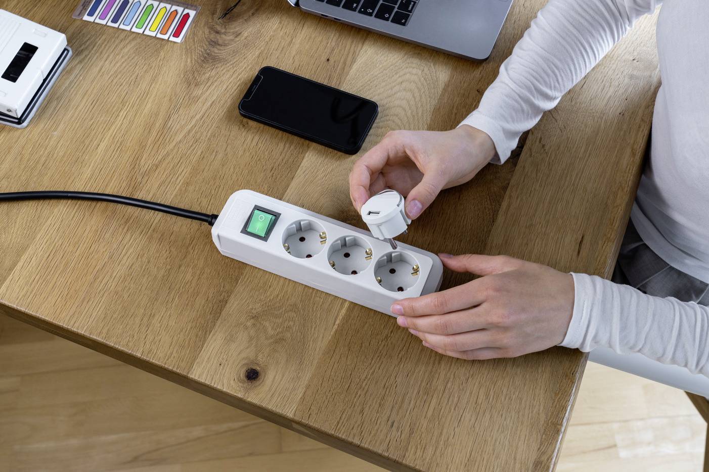 Person steckt einen Stecker in eine Steckdosenleiste auf einem Holztisch. Laptop und Smartphone sind daneben platziert.