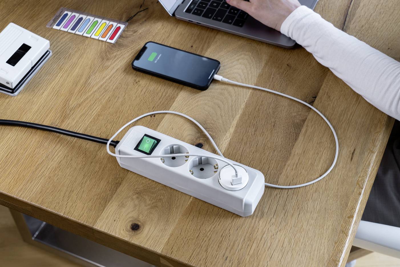 Eine Person arbeitet an einem Laptop auf einem Holztisch. Ein Smartphone wird über eine Mehrfachsteckdose geladen.