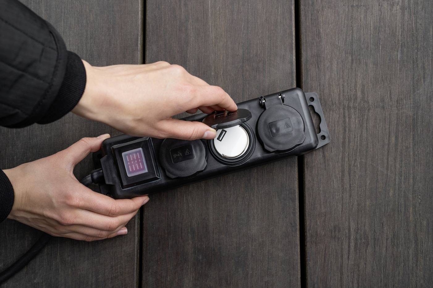 Eine Hand schließt das Schutzcover einer Steckdosenleiste im Freien. Die Leiste hat mehrere Steckplätze und ist gegen Feuchtigkeit geschützt.