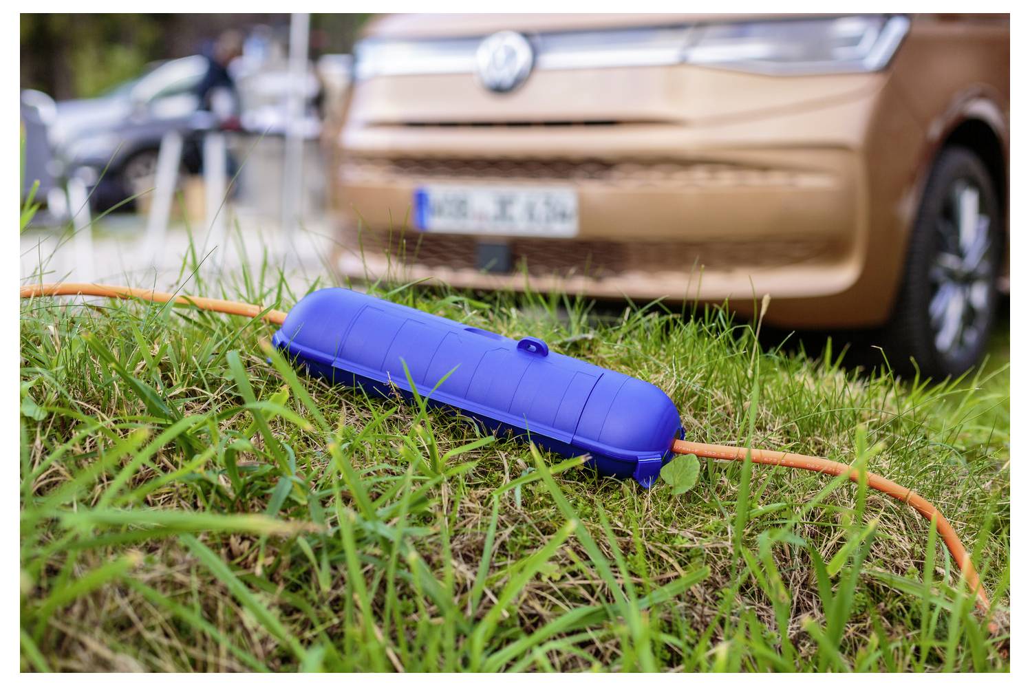 Blauer Ladestecker und oranges Kabel liegen im Gras vor einem geparkten Elektroauto.