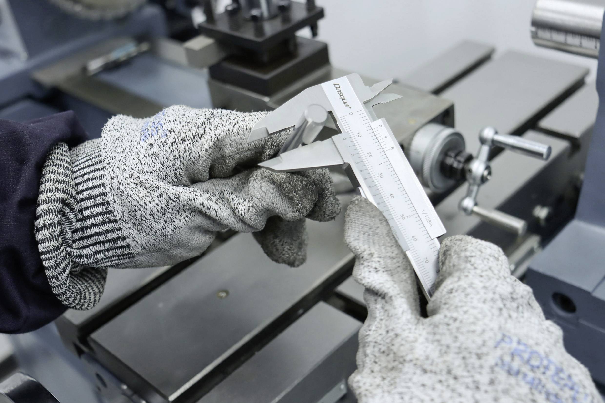 Eine Person mit grauen Handschuhen misst mit einem Messschieber ein Metallstück in einer Werkstattumgebung.