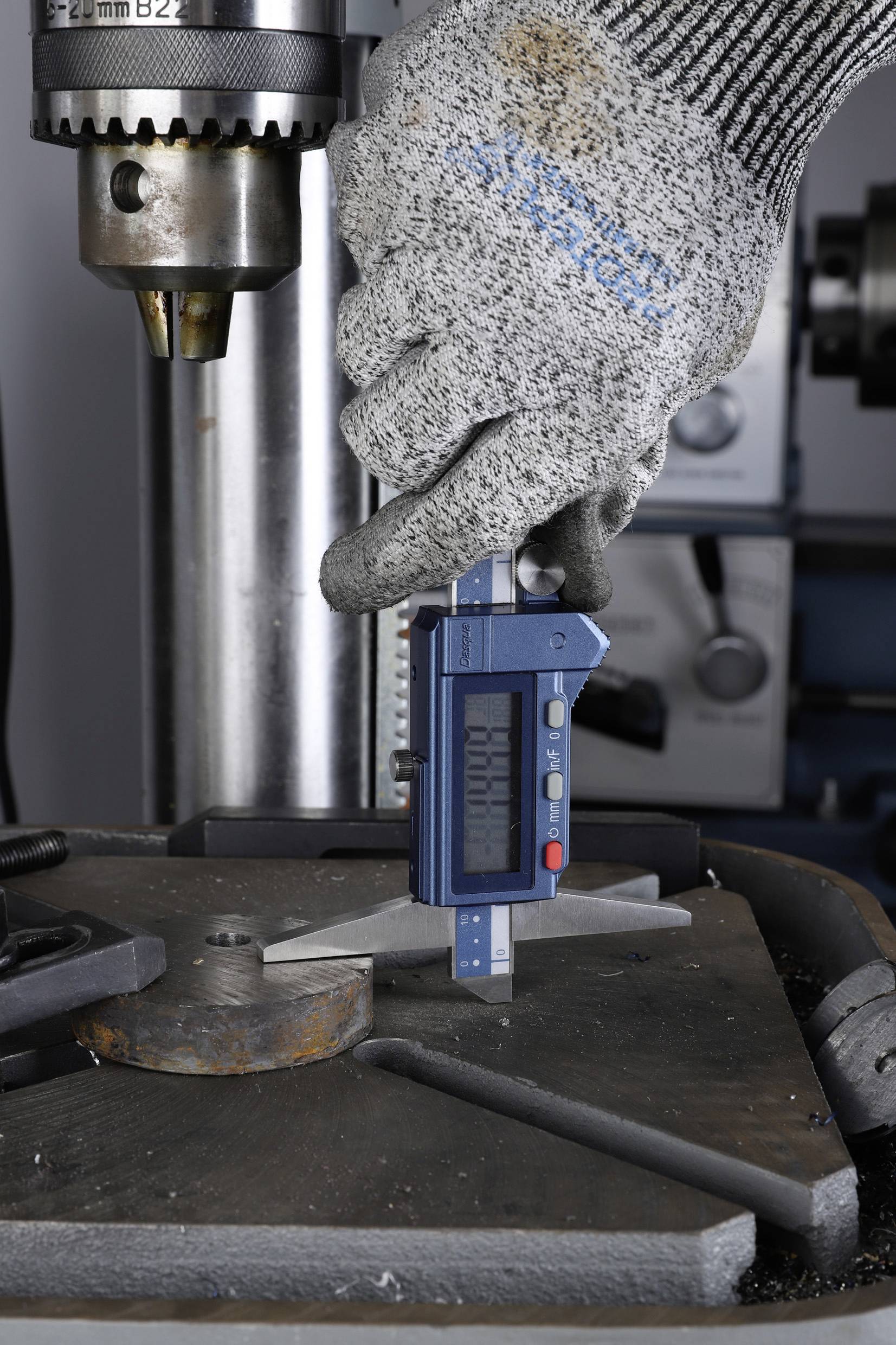 Eine behandschuhte Hand misst ein Metallteil auf einem Tisch mit einem digitalen Messschieber. Im Hintergrund befindet sich eine Bohrmaschine.