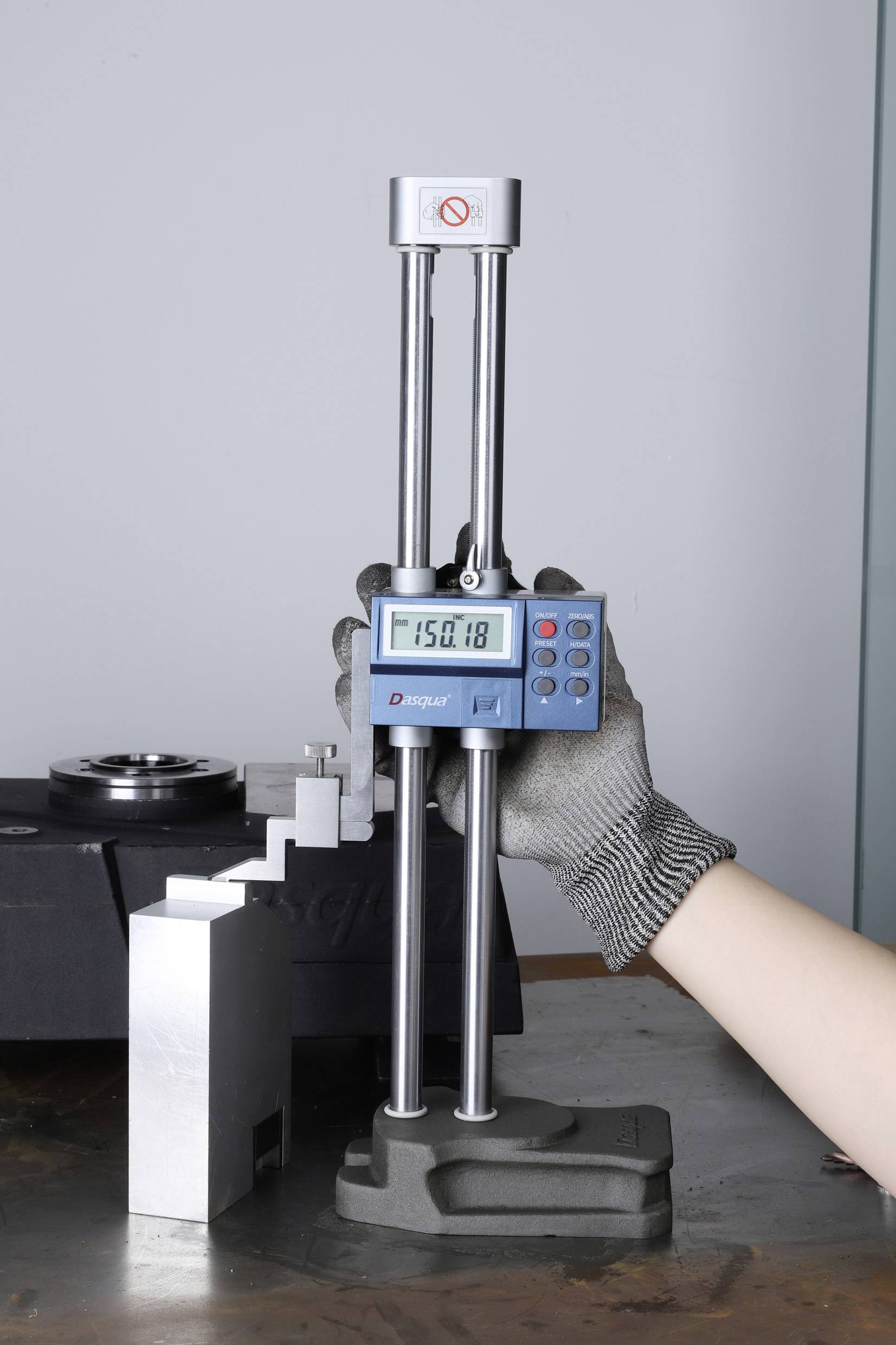 Digitale Höhenlehre misst Metallteil; Display zeigt '150.18'. Eine behandschuhte Hand hält das Gerät stabil.