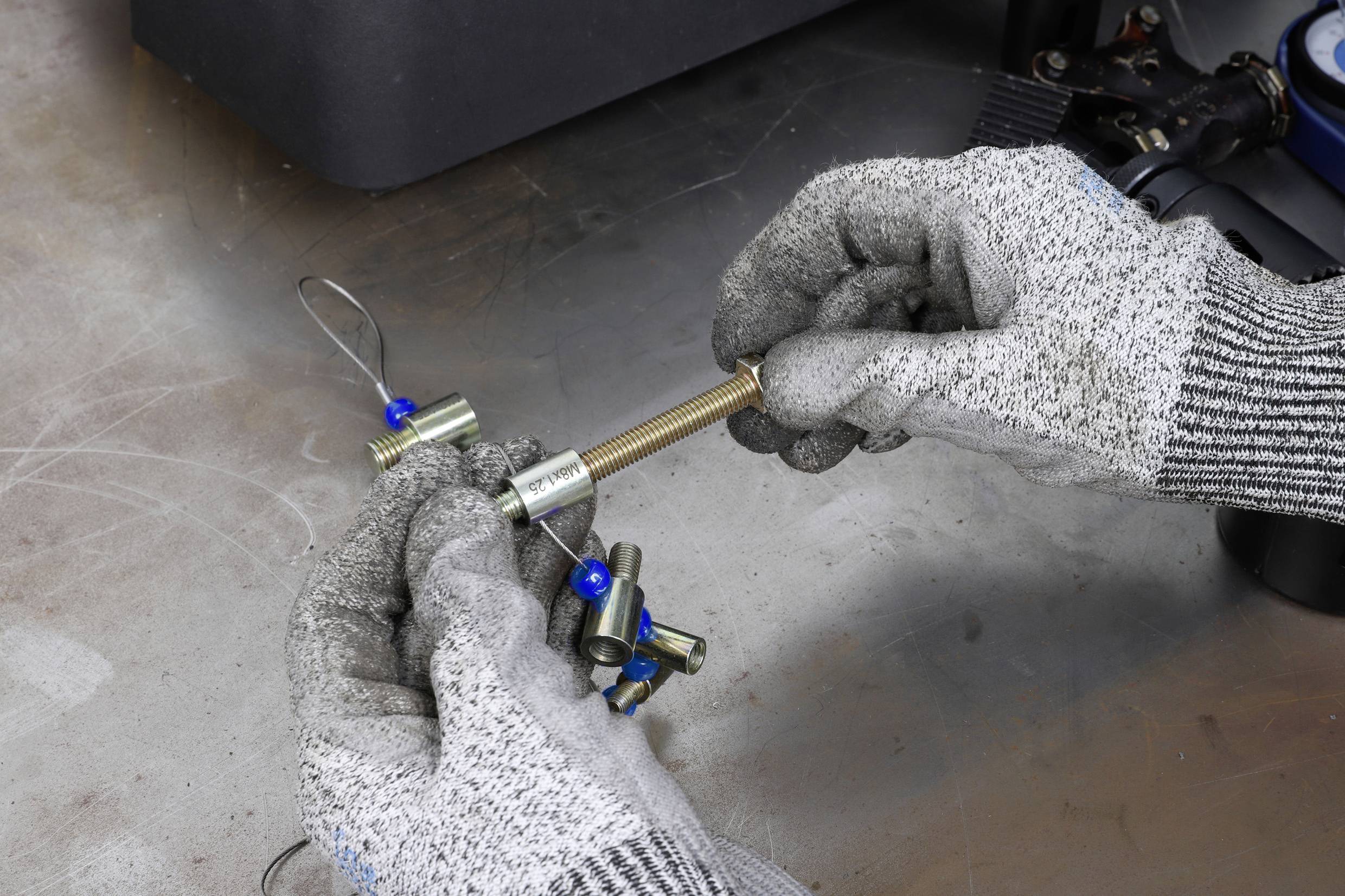 Person in grauen Handschuhen hält Drahtseilklemme mit Drahtseil, Metallschraube und Muttern. Arbeitsprozess an Werkbank.