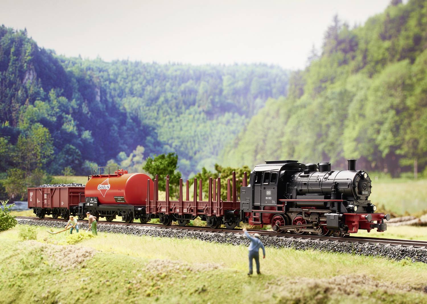 Eine Modelleisenbahn mit Dampflok zieht Güterwagen durch eine grüne Landschaft. Personenminiaturen arbeiten neben den Schienen.