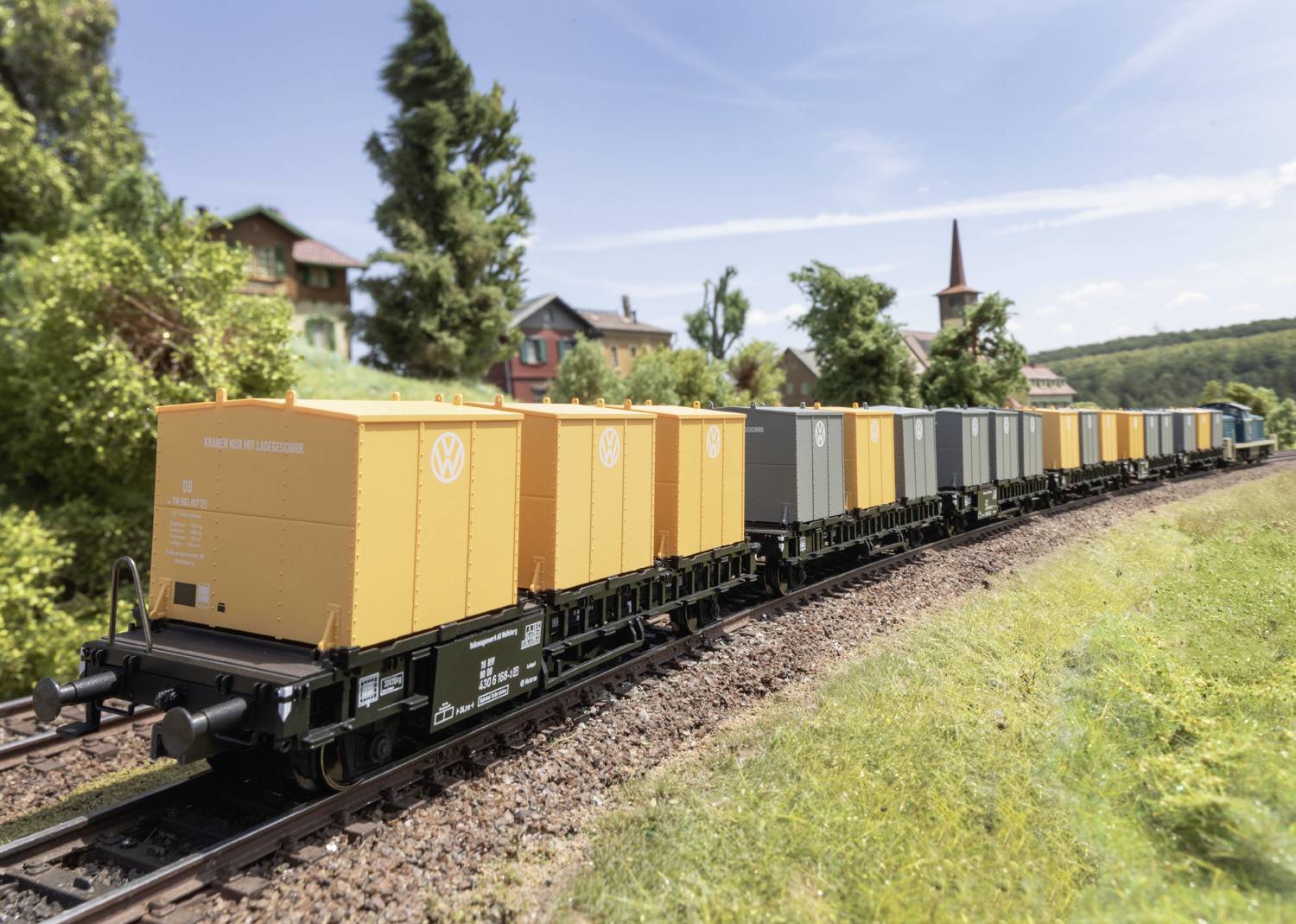 Eine Modelleisenbahn mit orangefarbenen und grauen Frachtcontainern fährt durch eine ländliche Landschaft bei klarem Himmel.