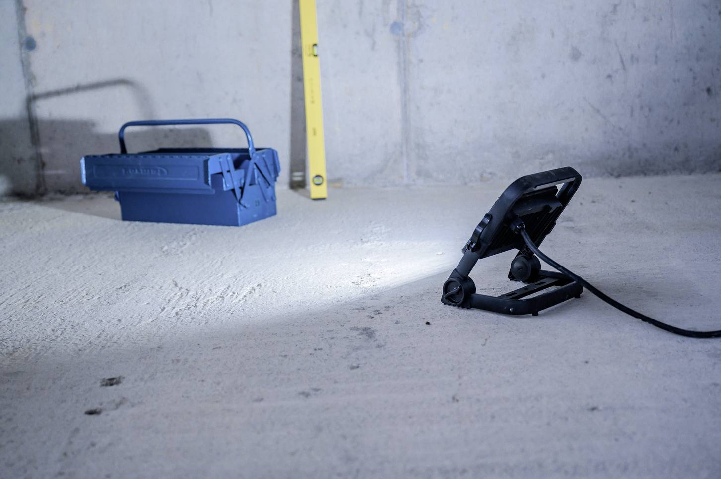 Ein blauer Werkzeugkasten steht auf einem Betonboden. Daneben befindet sich eine gelbe Wasserwaage. Im Vordergrund strahlt eine Lampe.