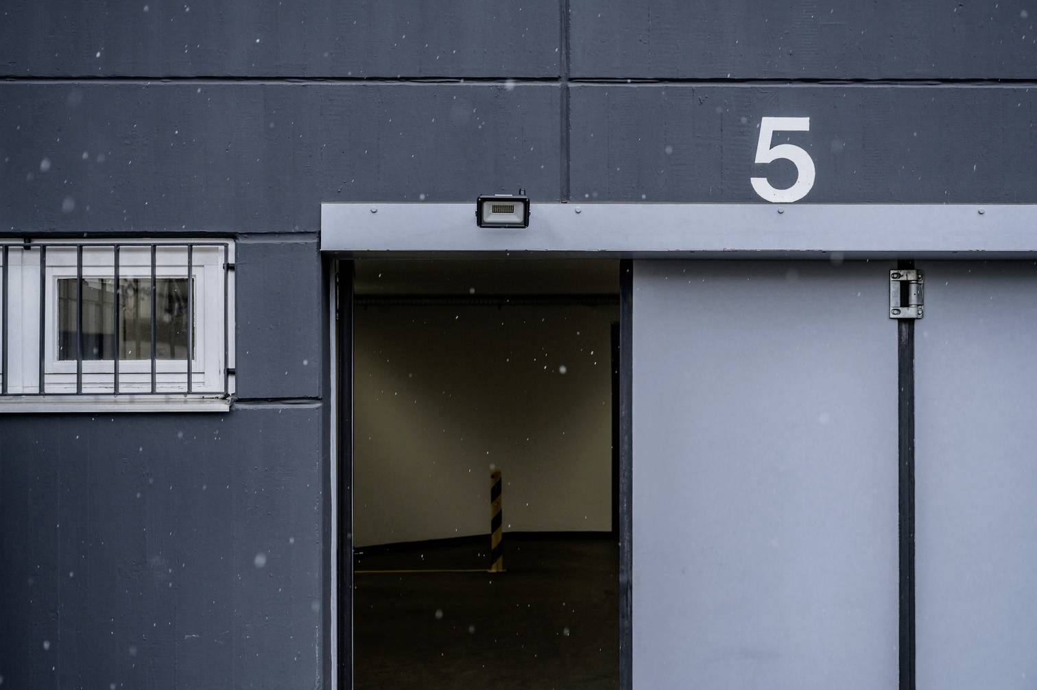 Offenes Garagentor mit der Nummer 5. Dunkler Innenbereich sichtbar, außen schneit es leicht. Ein schmaler Poller ist im Inneren erkennbar.