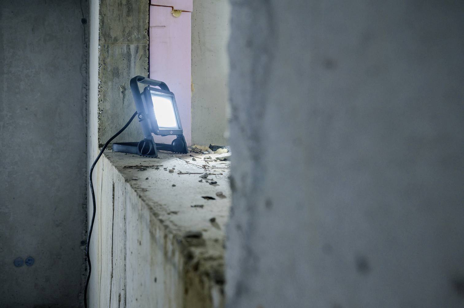 Ein Baustrahler steht auf einer staubigen Fensterbank in einem unvollendeten Raum. Das Licht erhellt die Betonwände.