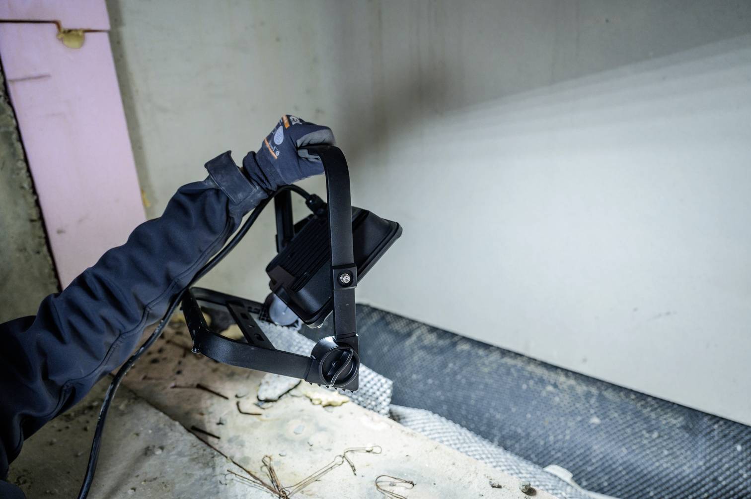 Eine Person mit Handschuhen hält einen Baustrahler, der auf eine Wand gerichtet ist. Der Ort scheint eine Baustelle zu sein.