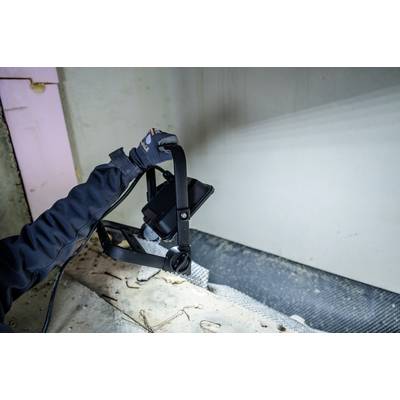 Eine Person mit Handschuhen hält einen Baustrahler, der auf eine Wand gerichtet ist. Der Ort scheint eine Baustelle zu sein.