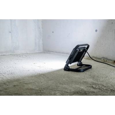 Ein tragbarer LED-Baustrahler leuchtet eine leere, graue Betonwand in einem minimalistisch gestalteten Raum aus.
