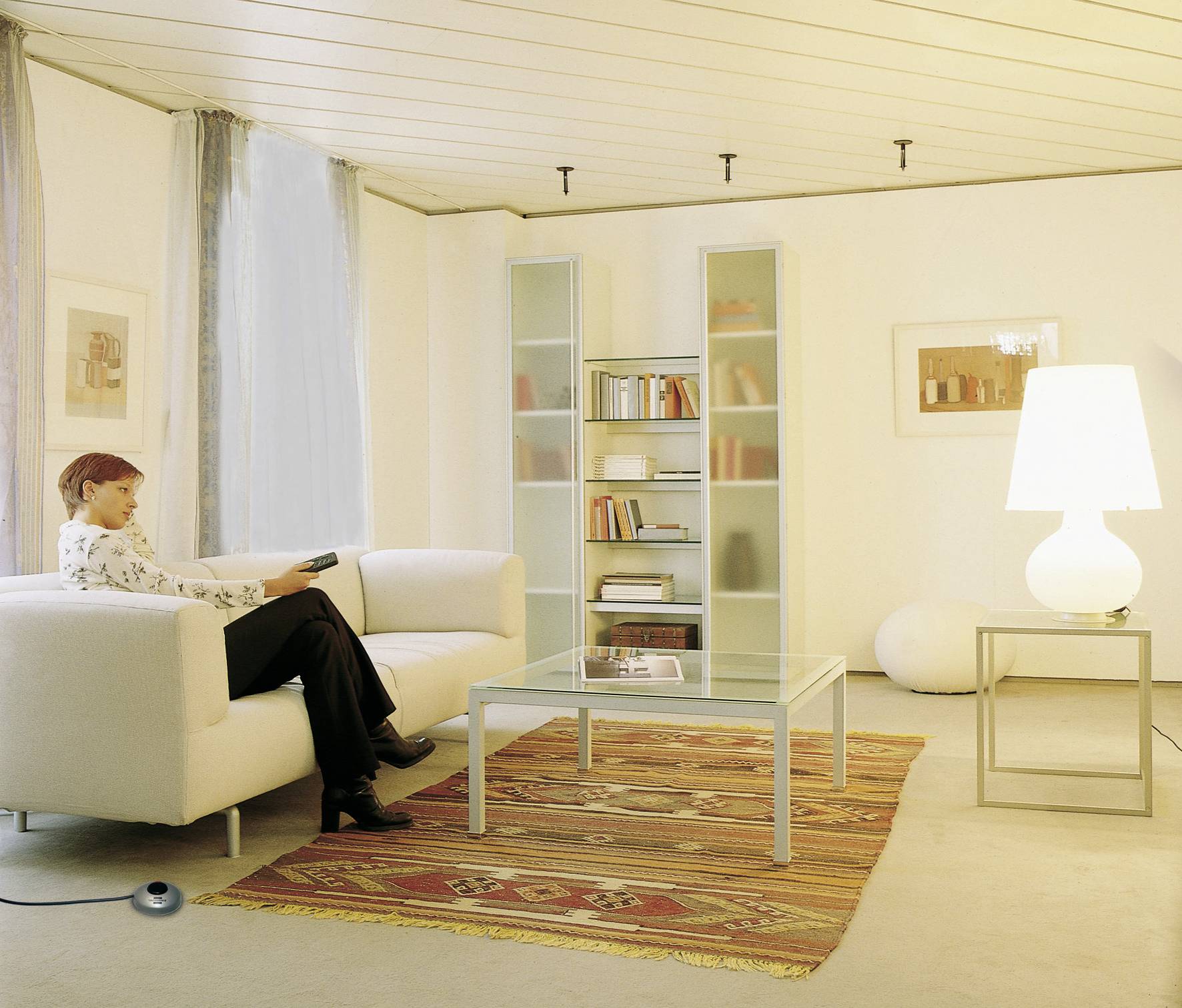 Eine Person sitzt auf einem Sofa in einem modernen Wohnzimmer mit hellem Interieur, Bücherregal und beleuchtetem Tisch.