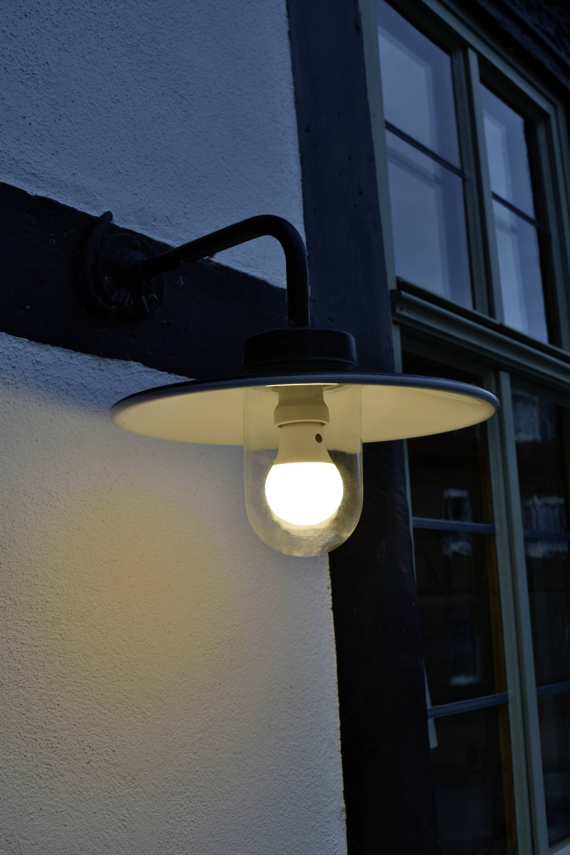 Außenlampe an einer Hauswand, beleuchtet in der Abenddämmerung, mit einem Fenster im Hintergrund.