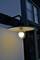 Eine eingeschaltete Außenlampe an einer weißen Wand neben einem Fenster, beleuchtet die Umgebung in der Dämmerung.