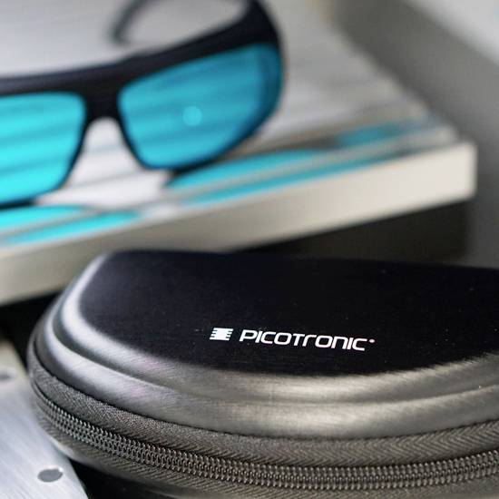 Brille mit blau getönten Gläsern neben einem Etui mit der Aufschrift 'picotronic'.