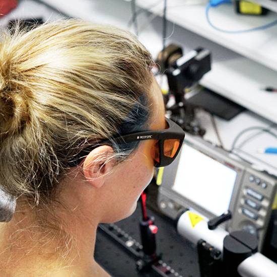 Eine Person, die mit Schutzbrille an einem wissenschaftlichen Laborgerät arbeitet. Im Hintergrund sind Geräte und Kabel sichtbar.