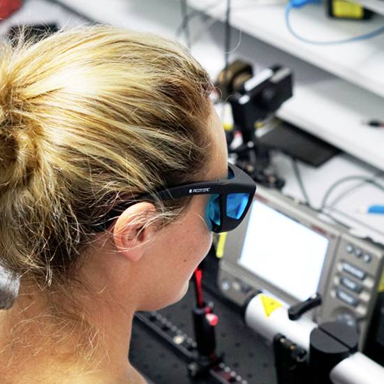 Eine Person mit Schutzbrille betrachtet ein technisches Gerät auf einem Labortisch, umgeben von wissenschaftlicher Ausrüstung.