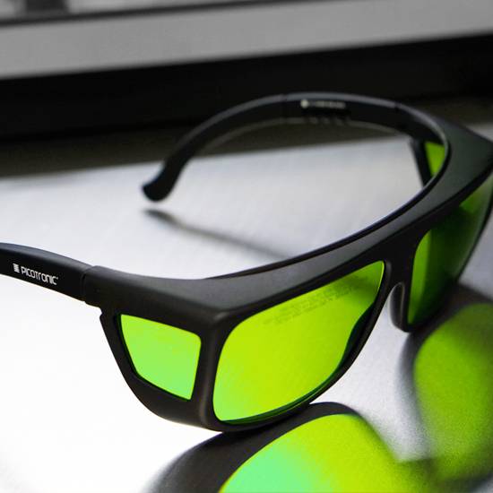 Schwarze Schutzbrille mit grünen Gläsern auf einer glänzenden Oberfläche, reflektiert Licht.