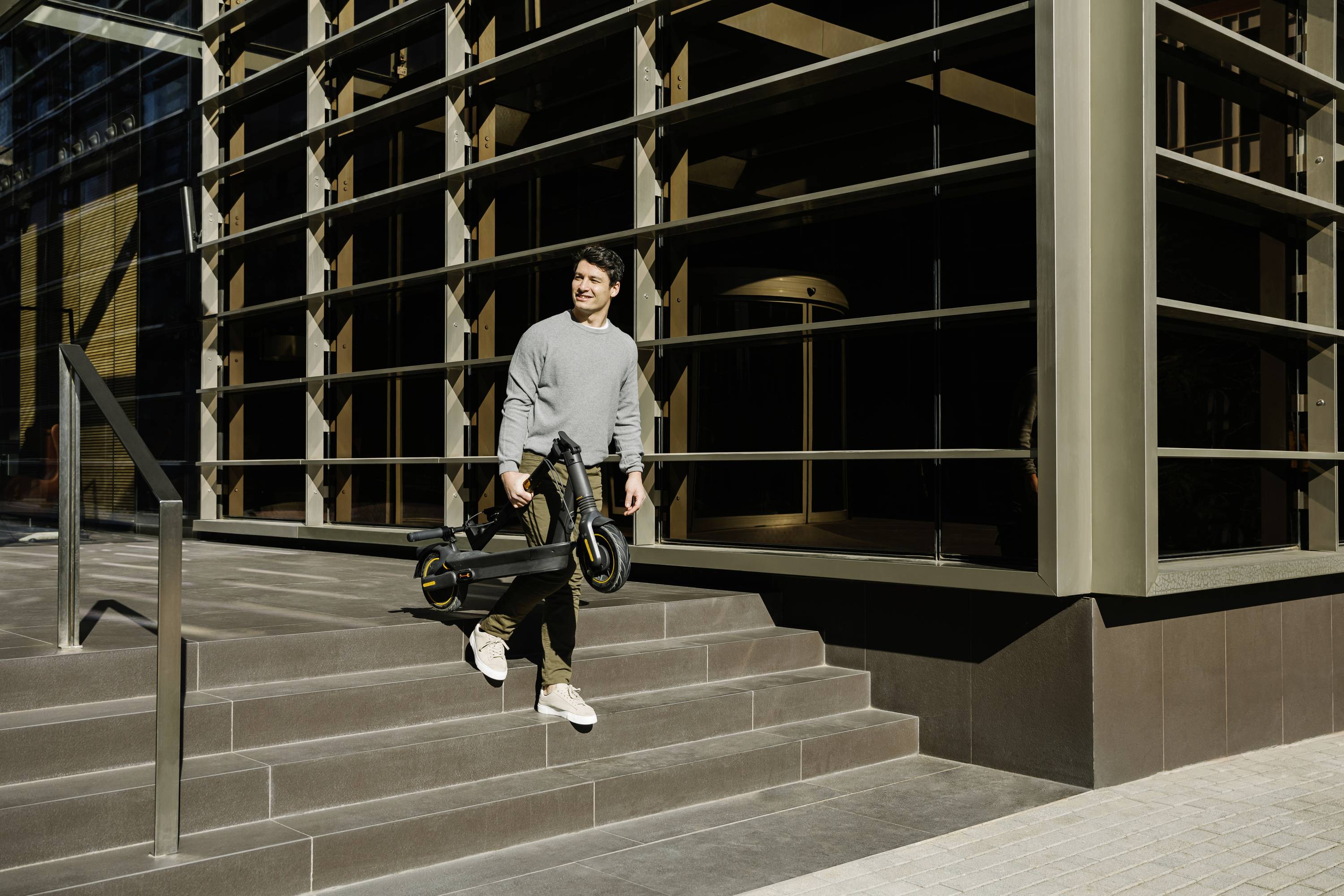 Person steht lächelnd auf Treppe vor modernem Gebäude und hält einen E-Scooter in der Hand.