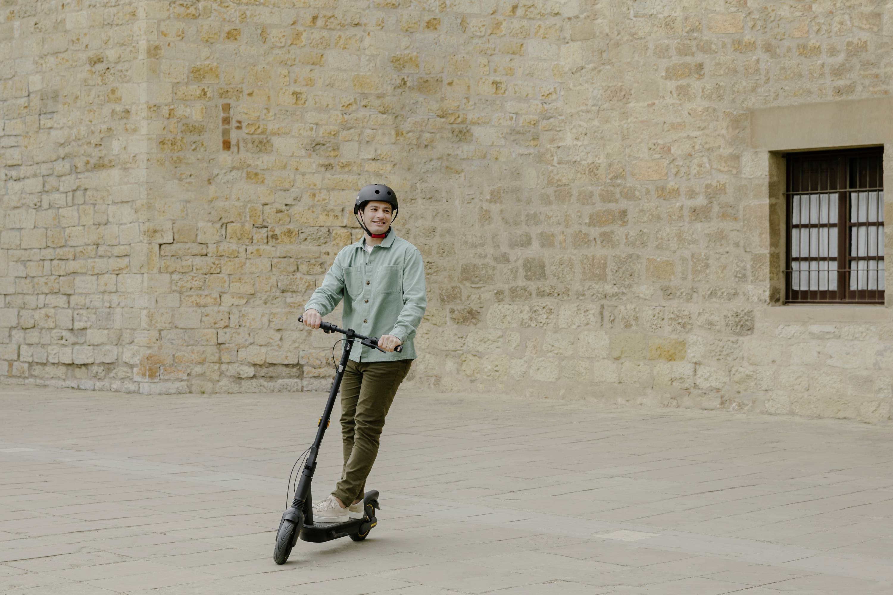 Eine Person fährt mit einem E-Scooter auf einem gepflasterten Platz vor einer alten Steinmauer. Sie trägt einen Helm und lächelt.