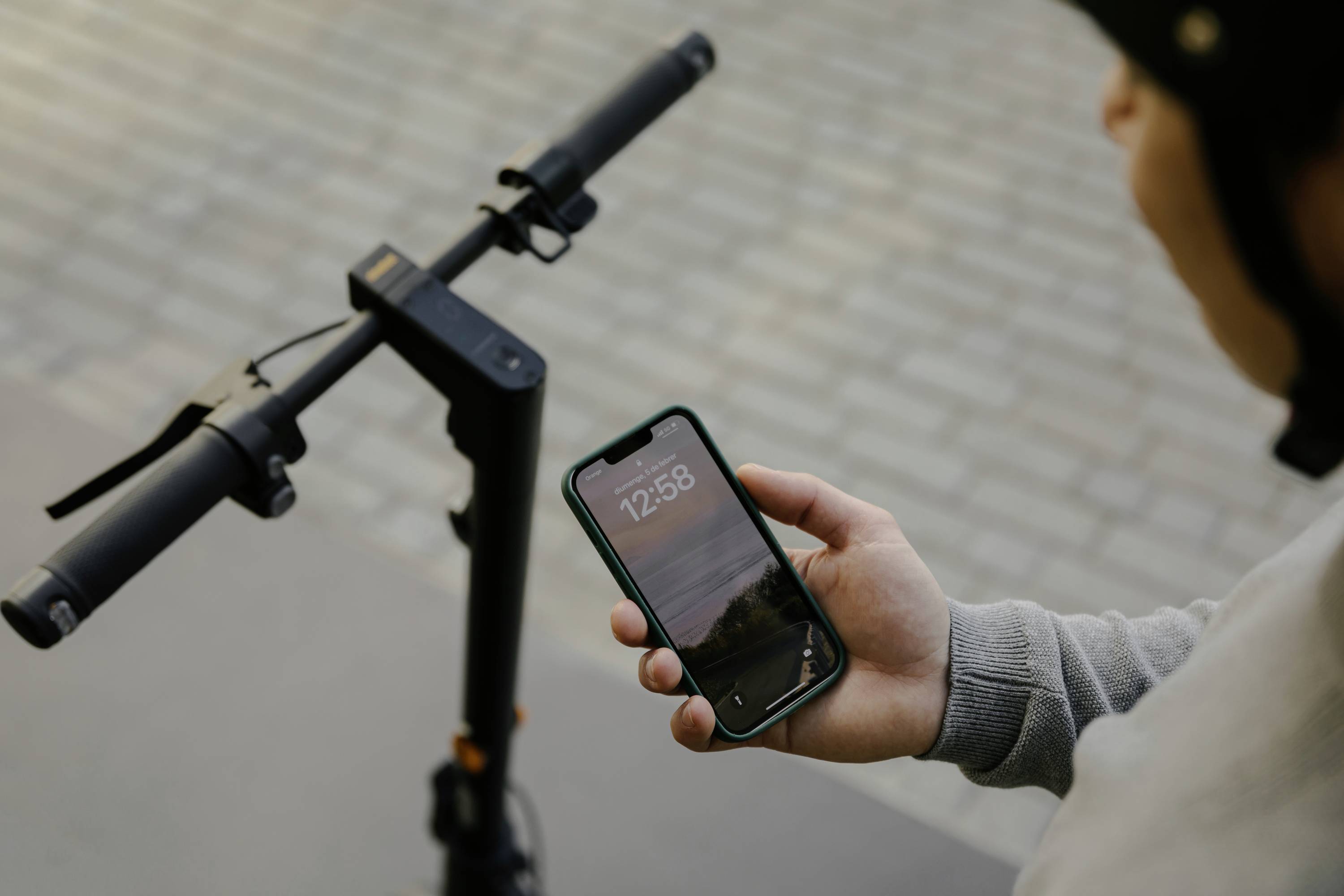 Person hält Smartphone mit Uhrzeit 12:58, neben einem E-Scooter-Lenker, auf Stadtstraße. Oberkörperansicht, von der Seite.