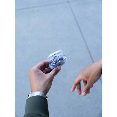 Zwei Hände in Anzügen; eine hält ein kleines, offenes, rundes Etui, das Ohrstöpsel enthält. Hintergrund ist ein grauer Boden.