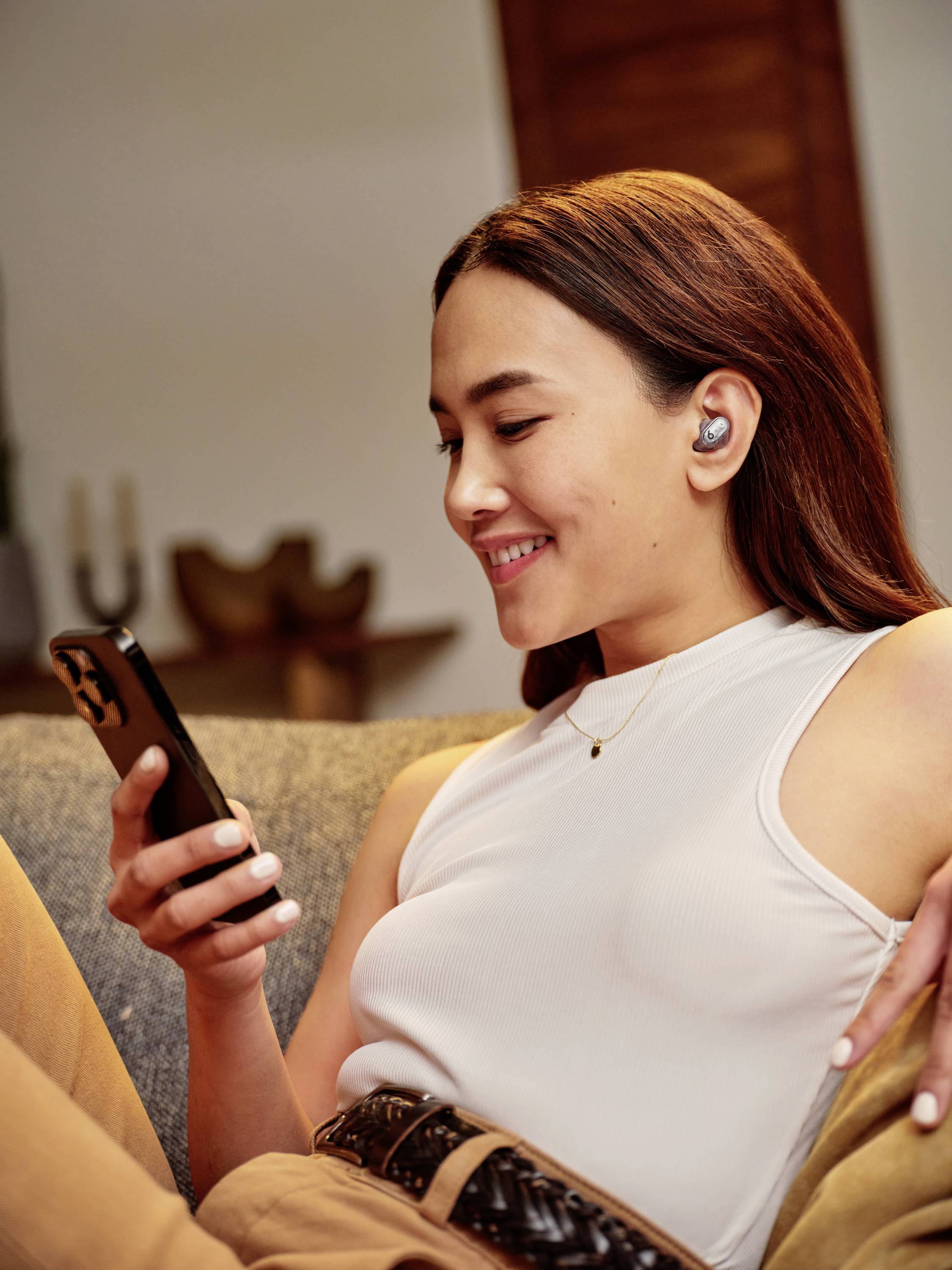 Eine Person sitzt entspannt auf einem Sofa, hört Musik mit kabellosen Ohrhörern und schaut lächelnd auf ein Smartphone.