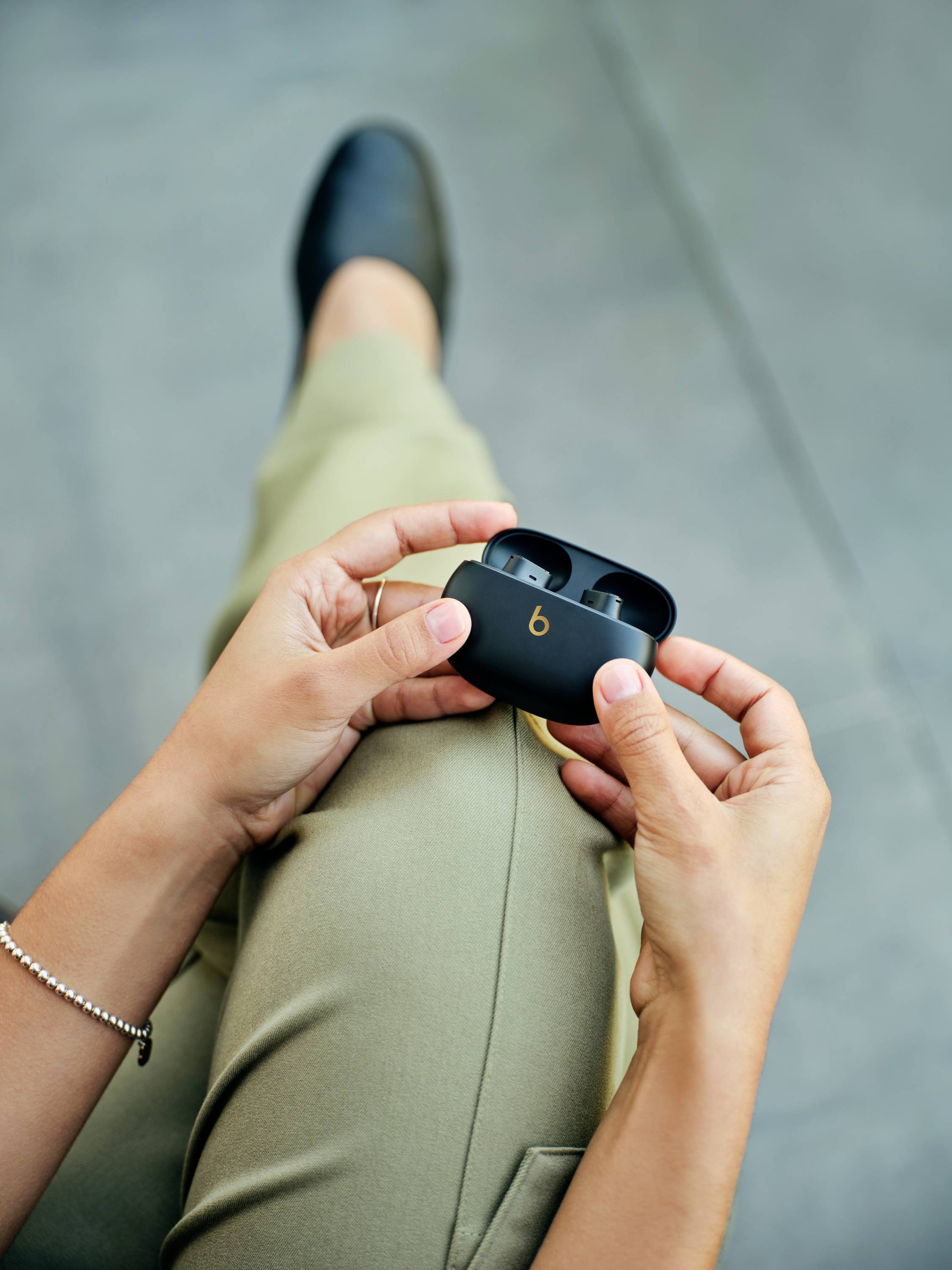 Person sitzt mit gekreuzten Beinen und hält schwarze kabellose Ohrhörer in einer Hand auf einem grauen Hintergrund.