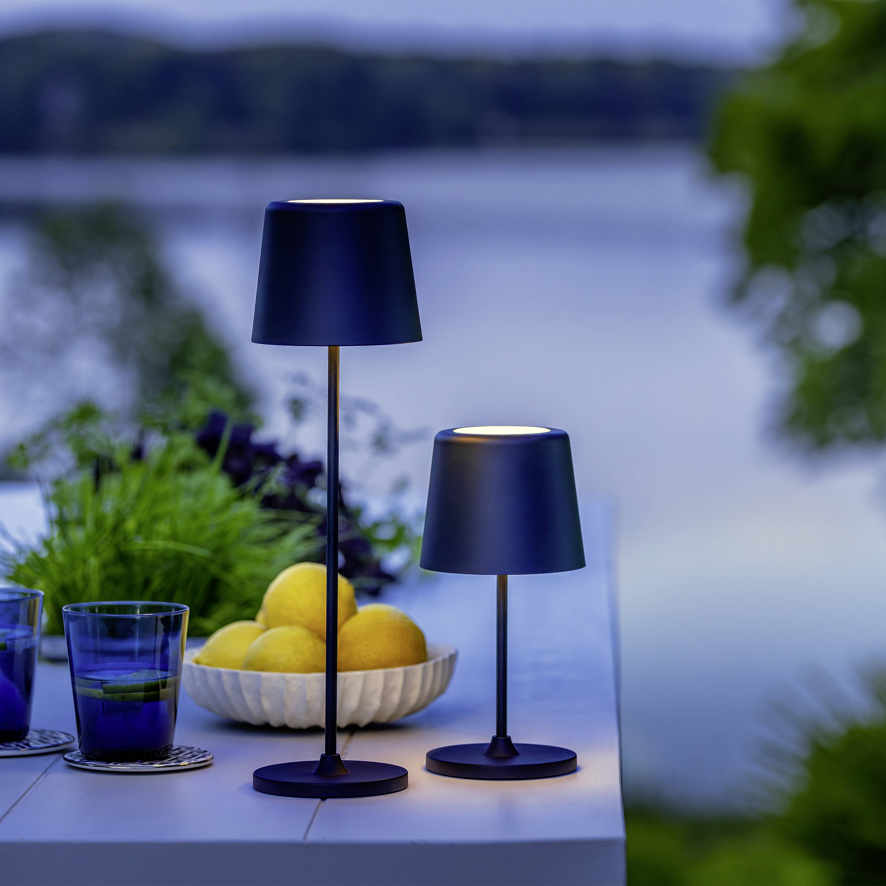 Ein Tisch im Freien mit zwei schwarzen Lampen, einem Schälchen Zitronen und drei blauen Gläsern, im Hintergrund ein verschwommener See.
