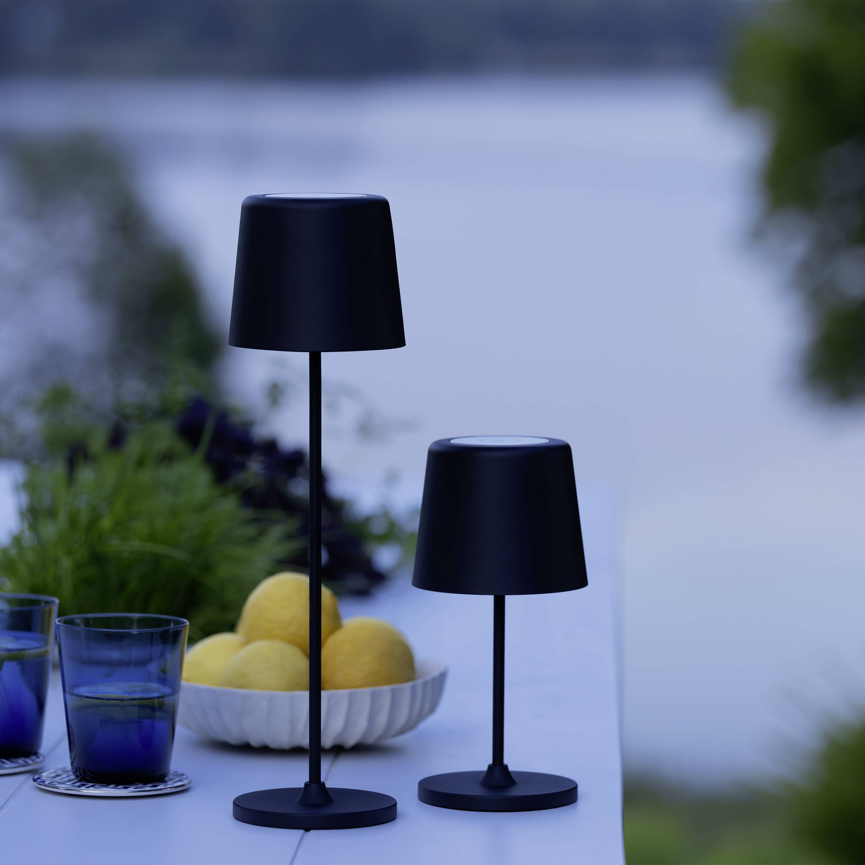 Zwei schwarze Tischlampen stehen auf einem weißen Tisch neben Gläsern und Zitronen. Im Hintergrund ist ein unscharfer See zu sehen.