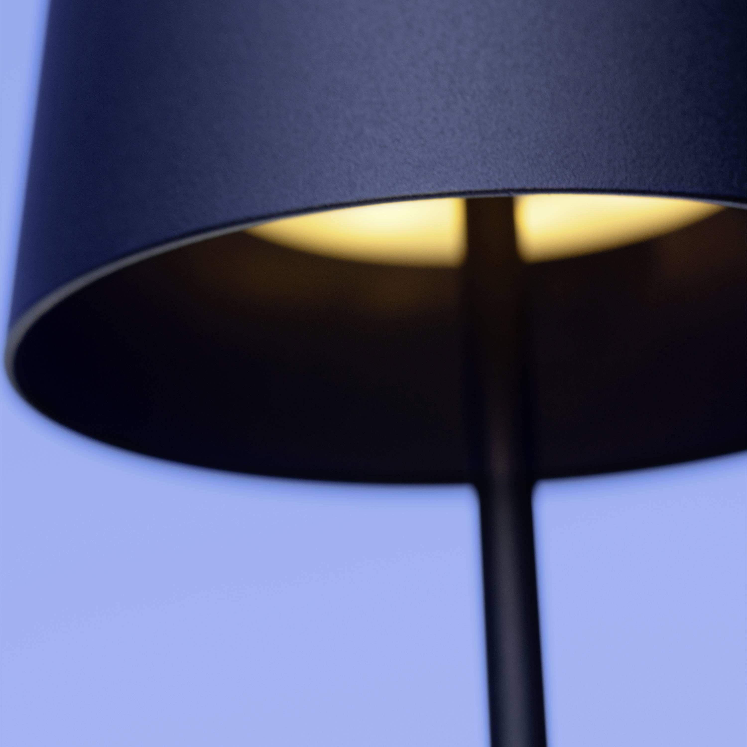 Nahaufnahme einer leuchtenden Tischlampe mit dunkelblauem Schirm vor einem hellblauen Hintergrund.