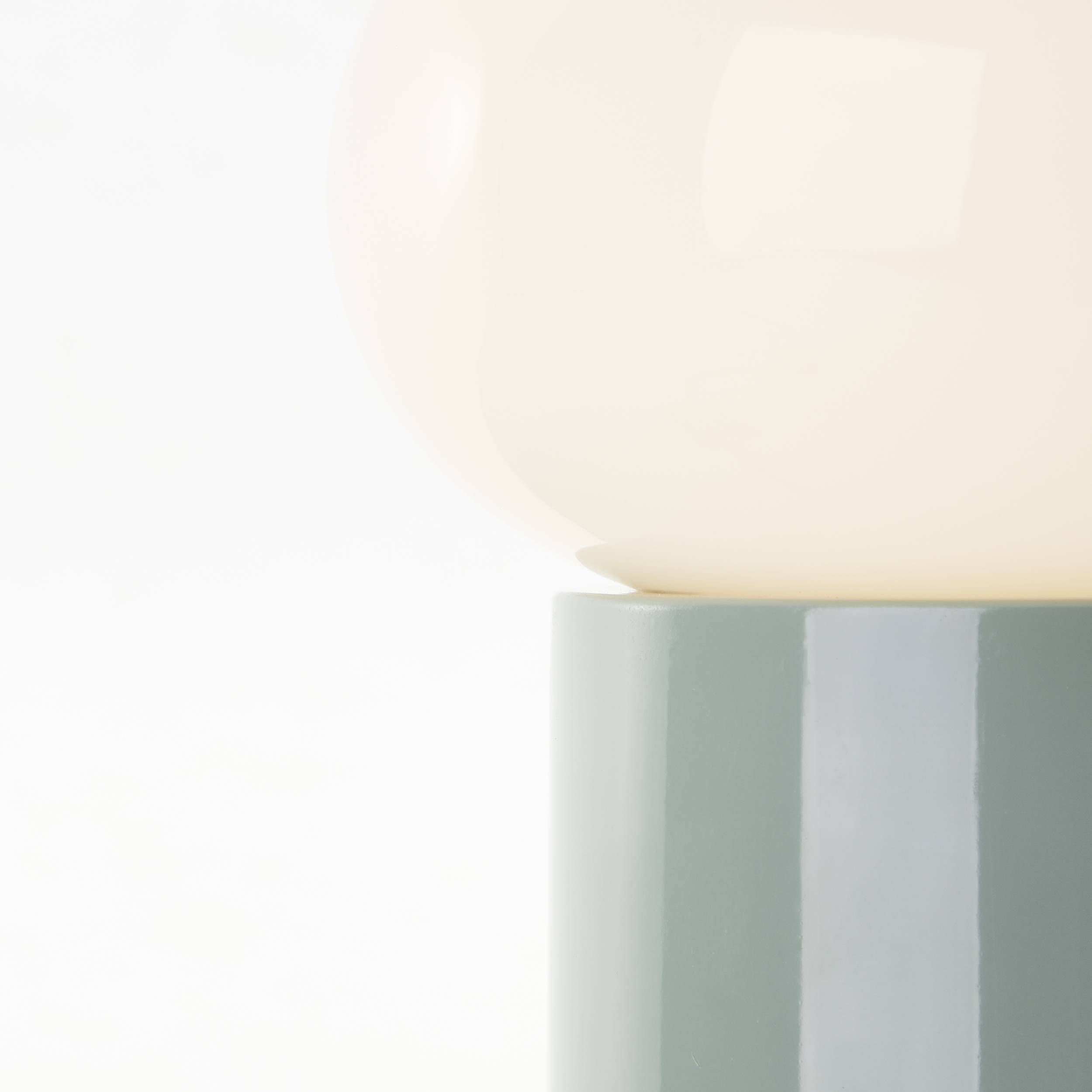 Eine minimalistische Lampe mit rundem, cremefarbenem Schirm auf grauem, glänzendem Sockel vor weißem Hintergrund.