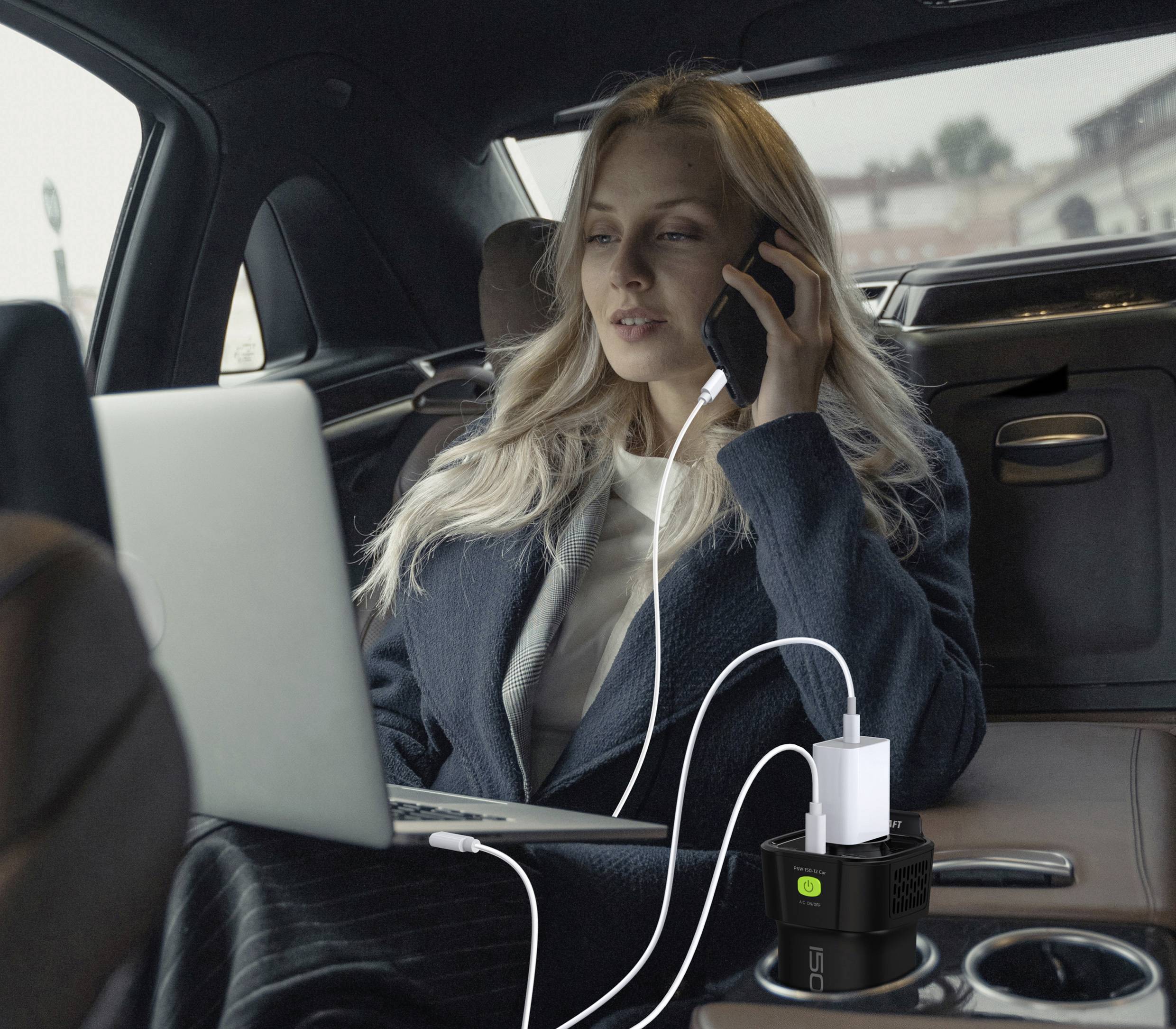 Eine Frau telefoniert und arbeitet auf einem Laptop in einem Auto. Mehrere Geräte sind über ein Ladegerät auf der Mittelkonsole angeschlossen.