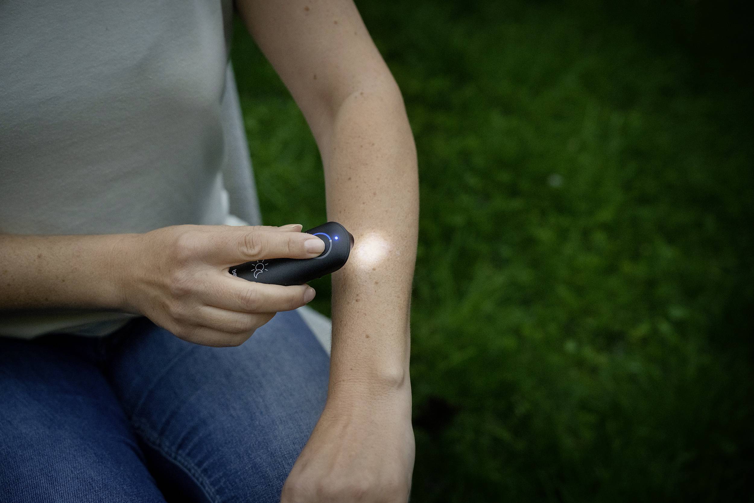 Eine Person hält ein tragbares Hautuntersuchungsgerät an ihren Arm, während sie draußen auf Gras sitzt.