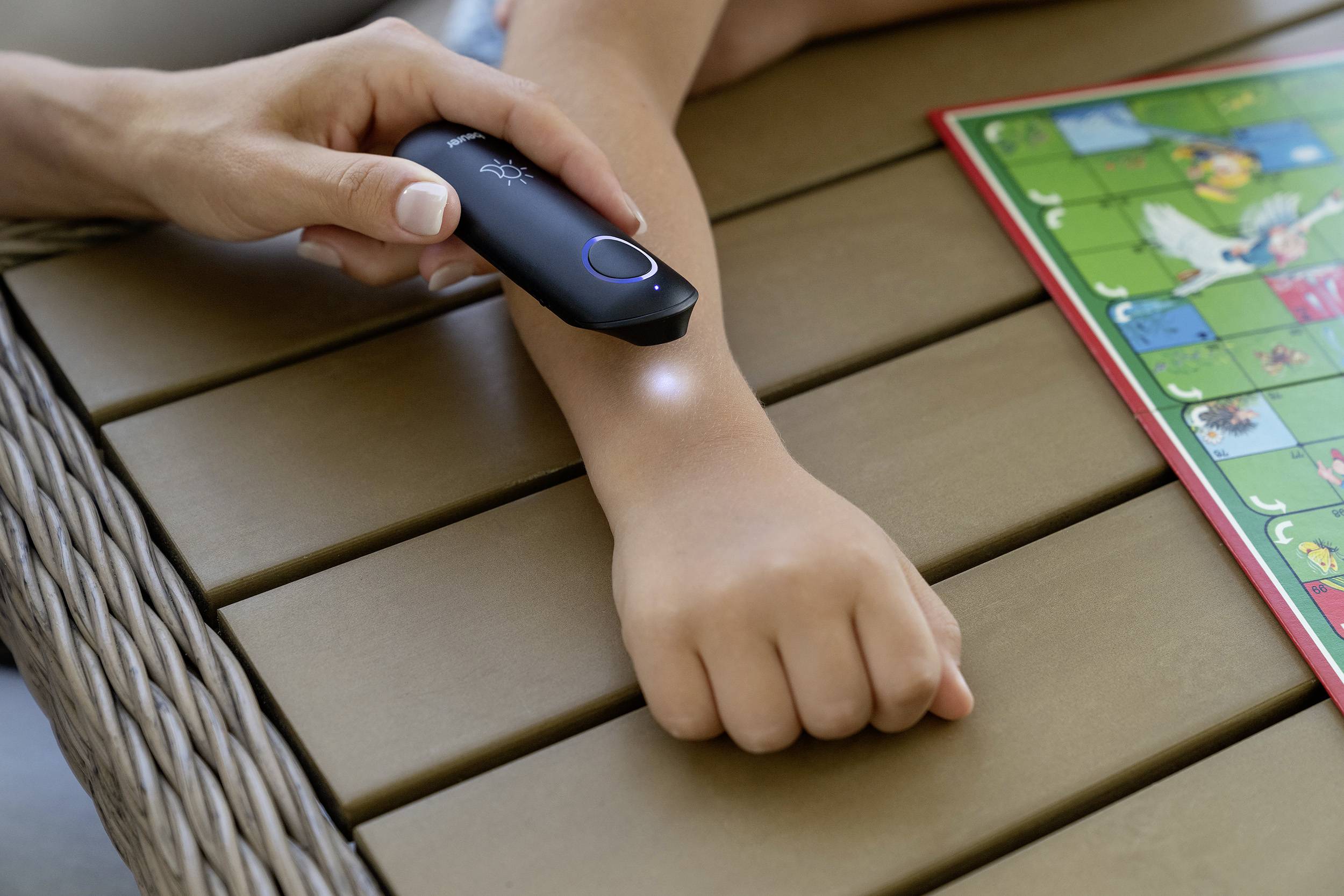 Eine Hand scannt mit einem tragbaren Gerät den Arm eines Kindes, das auf einem Tisch liegt. Daneben liegt ein buntes Spielfeld.