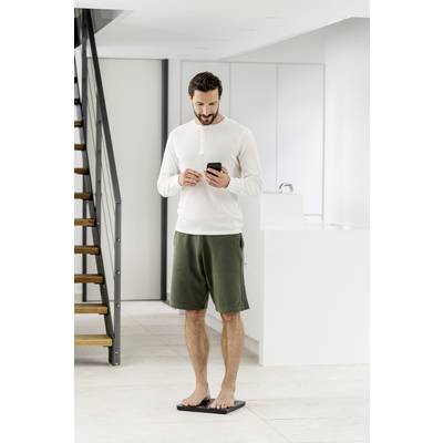 Ein Mann steht auf einer Waage in einem modernen, hellen Raum und schaut auf sein Smartphone.