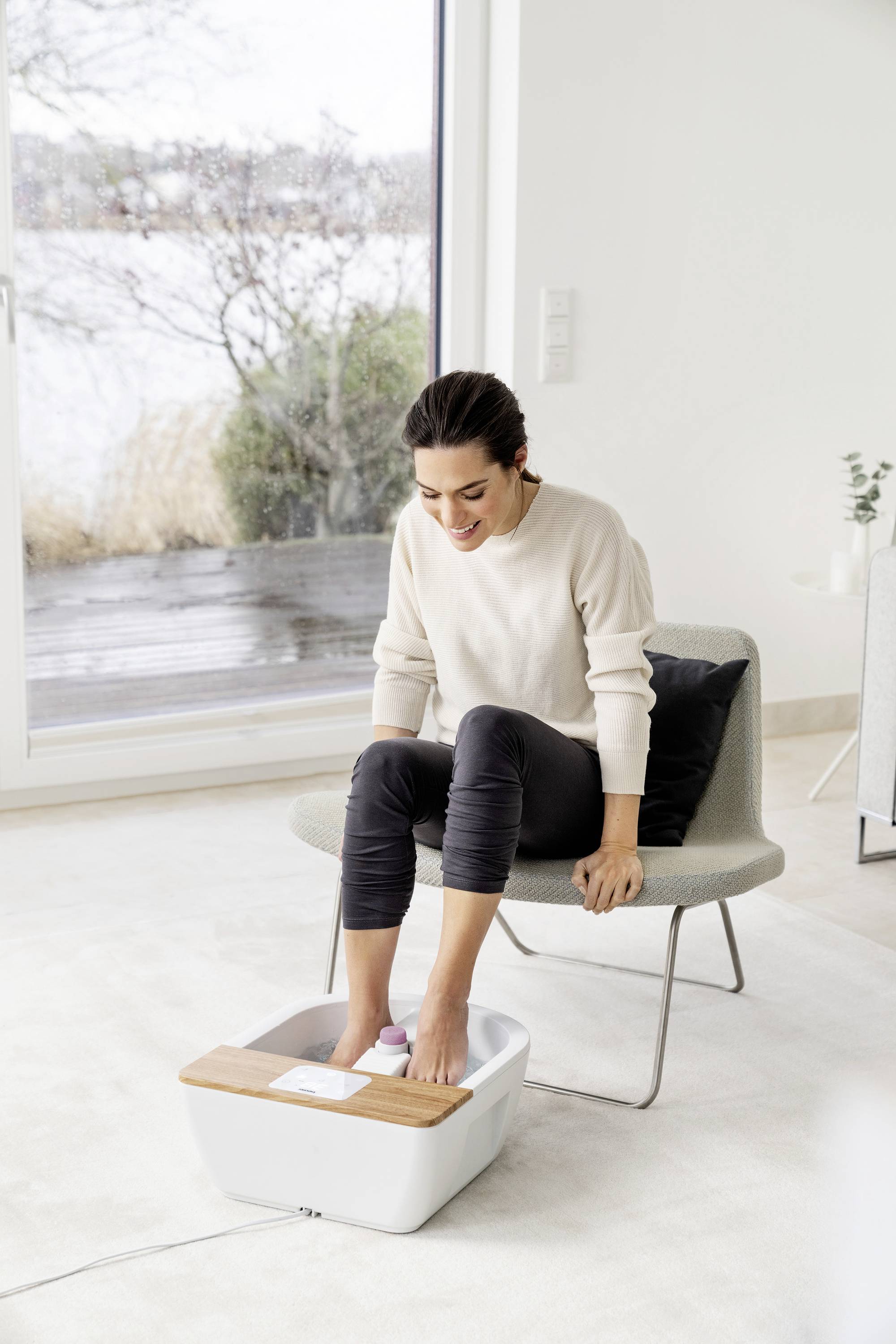 Eine Frau sitzt auf einem Stuhl und benutzt ein Fußbad in einem hellen Raum mit großen Fenstern, die eine Winterlandschaft zeigen.