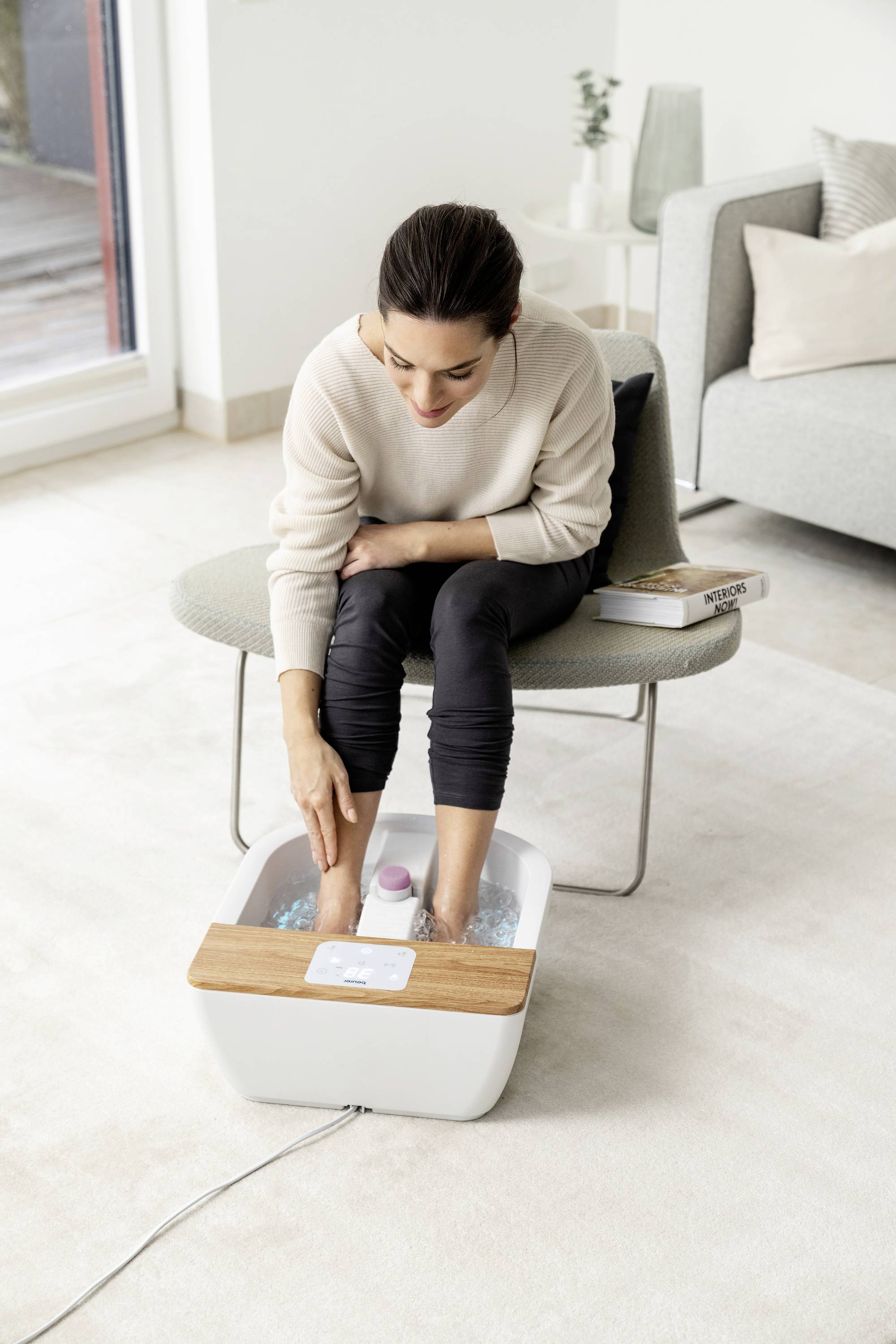 Eine Frau sitzt in einem modernen Wohnzimmer auf einem Stuhl und genießt ein Fußbad mit einem Holzdeckel-Fußsprudelbad.