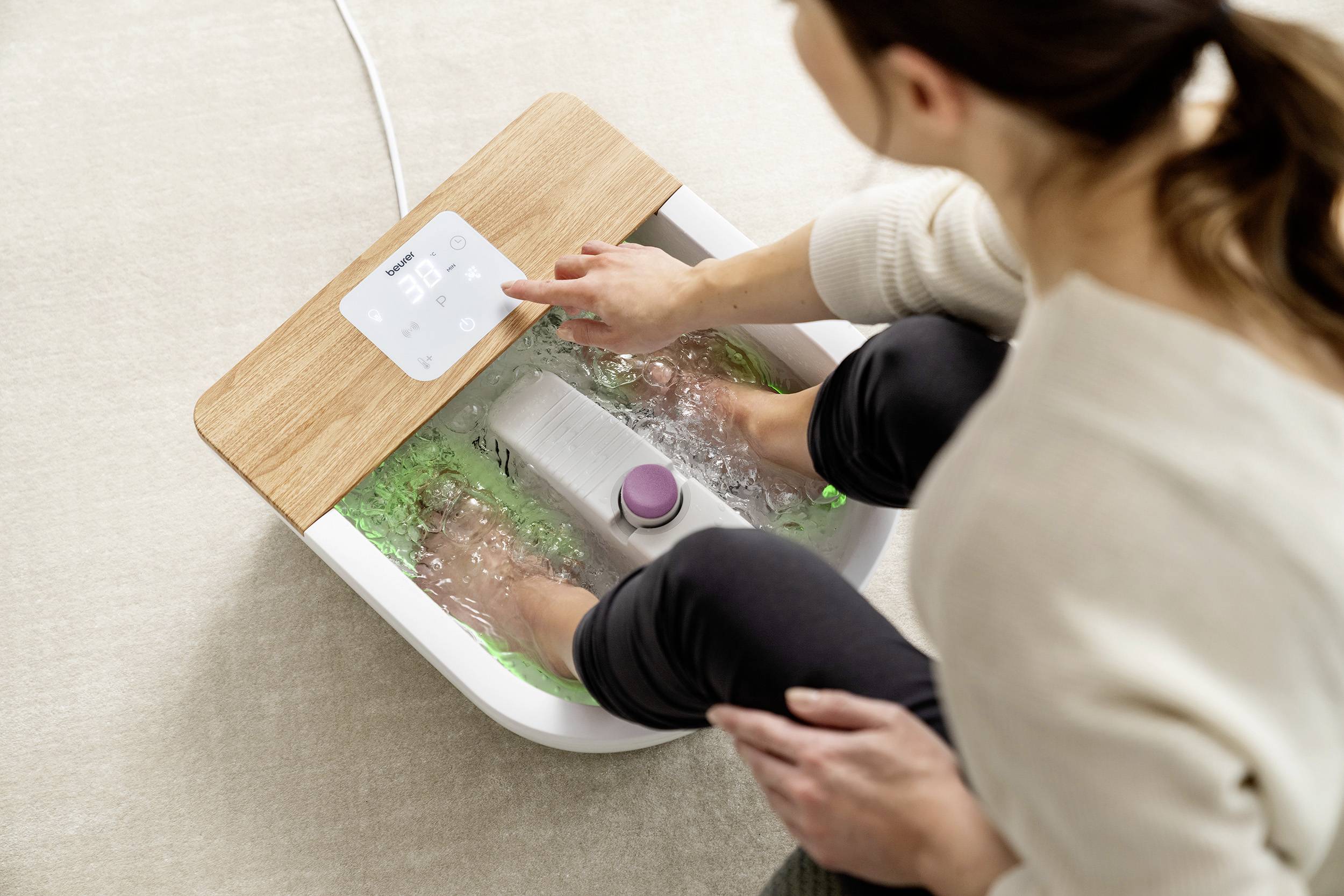 Eine Frau nutzt ein elektrisches Fußbad mit Sprudelfunktion; sie drückt auf das Bedienfeld. Entspannte häusliche Umgebung.