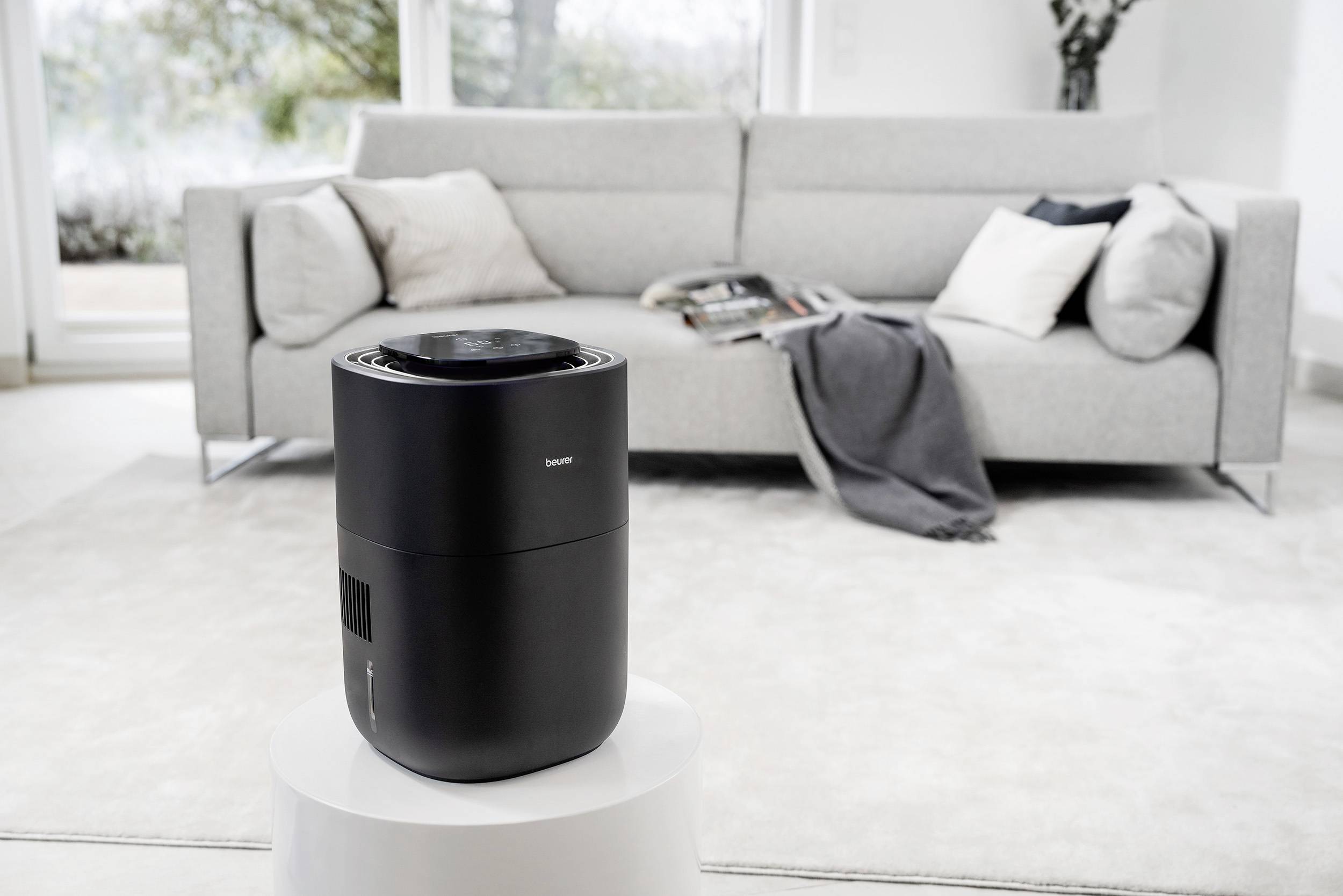 Ein schwarzer Luftreiniger steht auf einem weißen Tisch in einem modernen Wohnzimmer mit grauem Sofa und Kissen im Hintergrund.