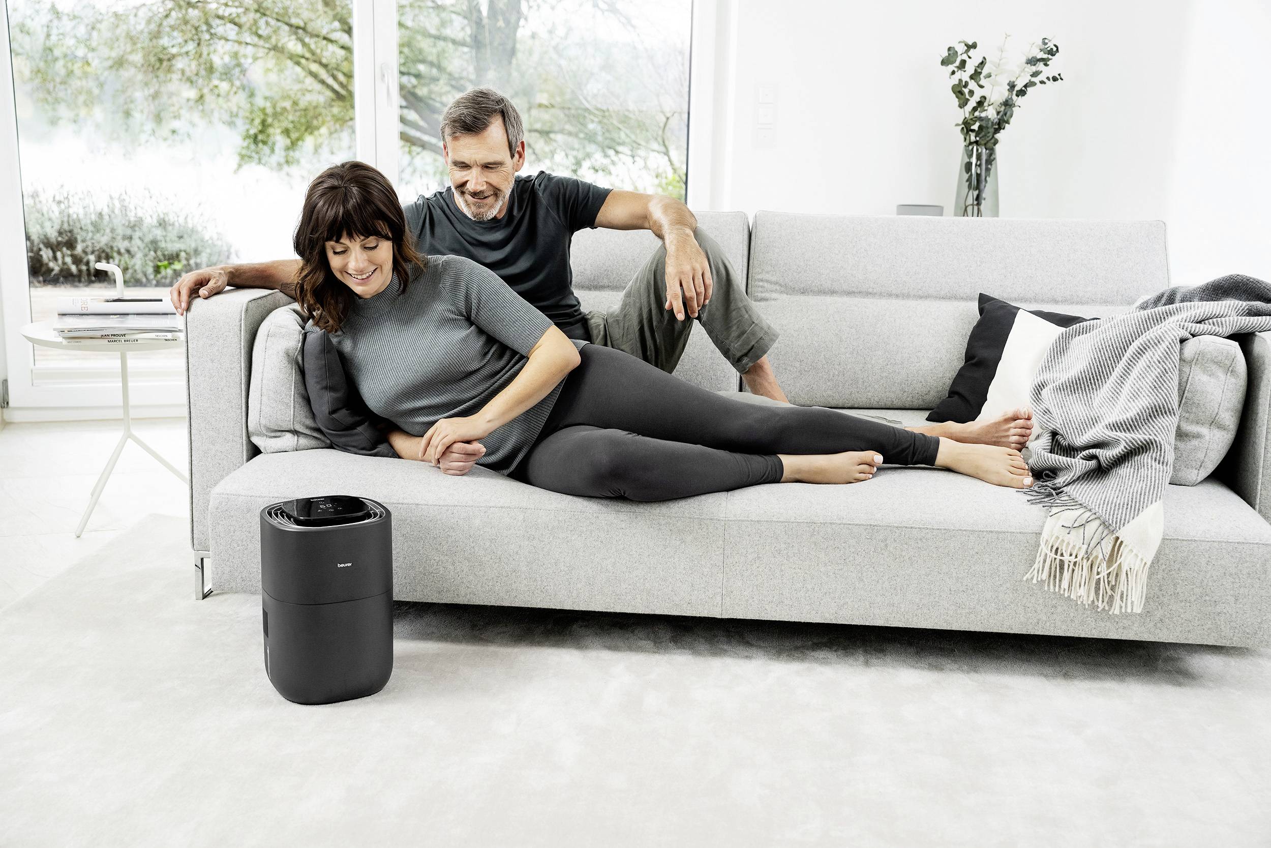 Ein Paar entspannt auf einem grauen Sofa in einem modernen, hellen Raum. Ein Luftreiniger steht davor. Ein großes Fenster zeigt eine grüne Landschaft.