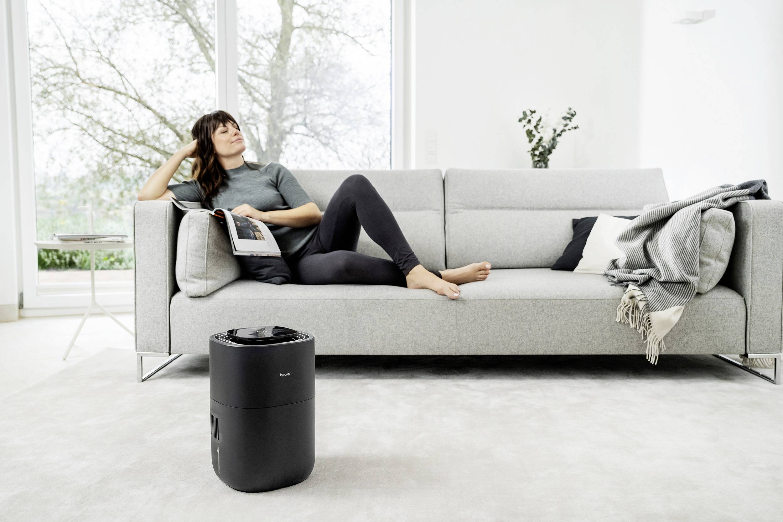 Frau liest auf einem Sofa in einem modernen Wohnzimmer, ein schwarzer Luftreiniger steht auf dem Boden.