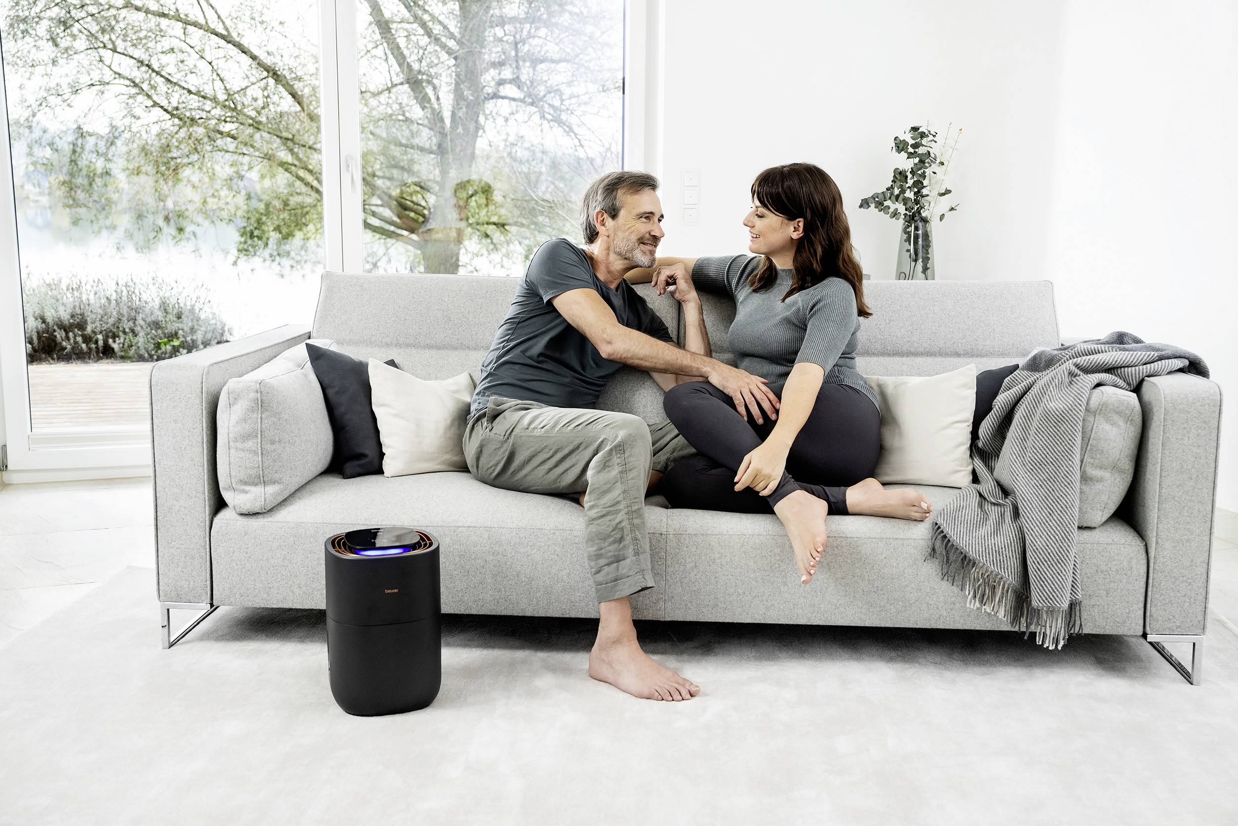 Ein Paar sitzt auf einem Sofa in einem modernen Wohnzimmer. Sie unterhalten sich entspannt. Neben dem Sofa steht ein Luftreiniger.