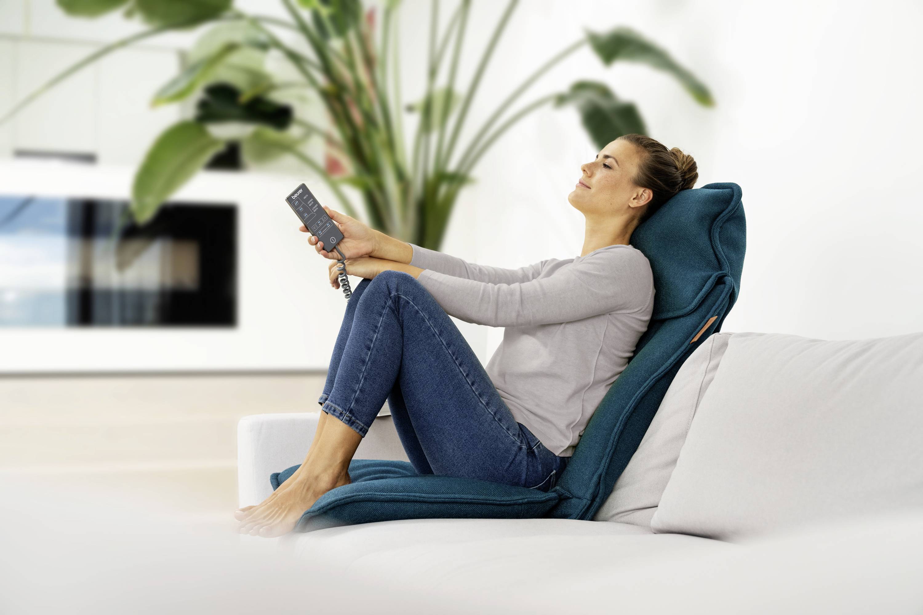 Frau sitzt entspannt auf einem Sofa mit einer Fernbedienung in der Hand, umgeben von Pflanzen im Hintergrund.