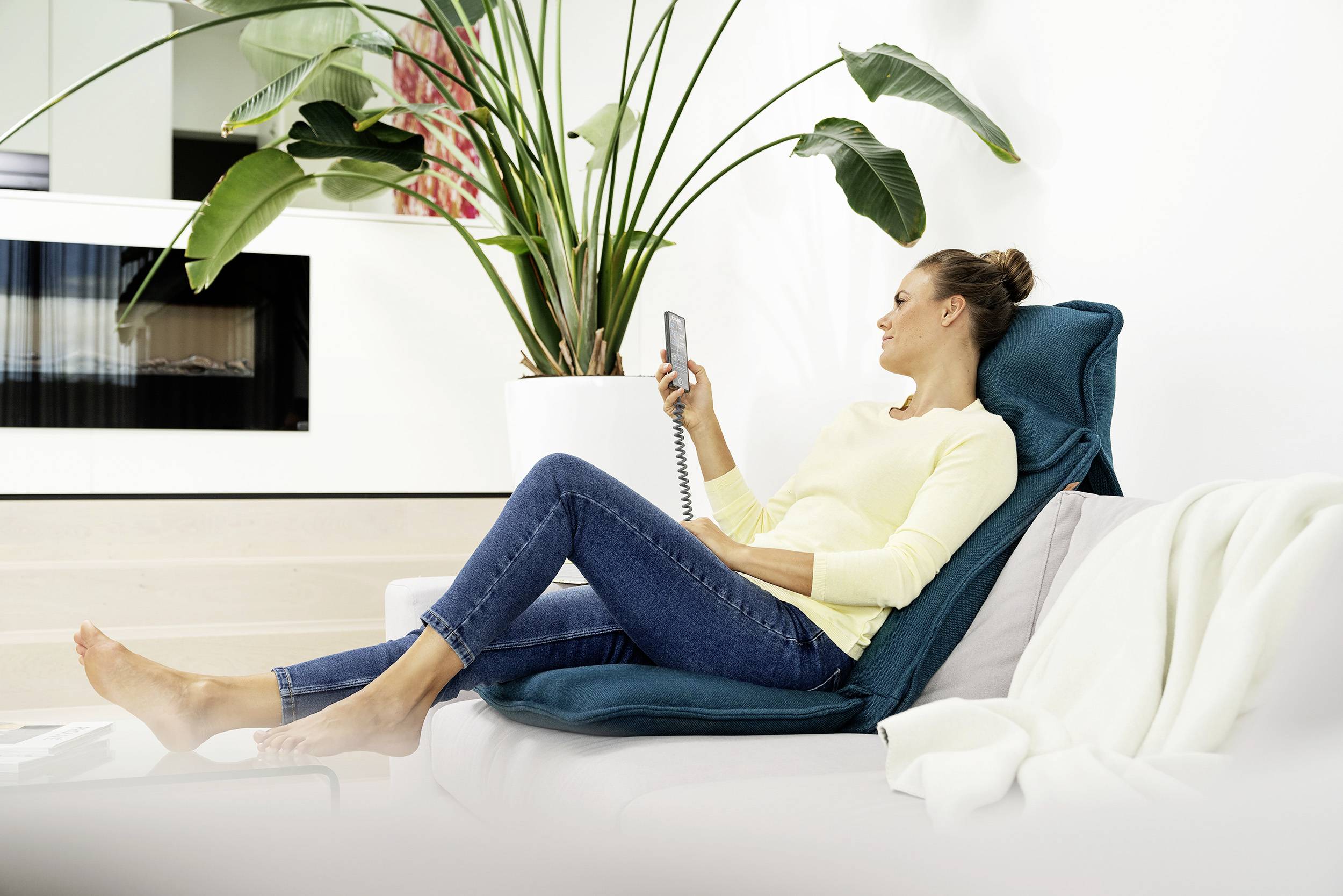 Eine Frau sitzt entspannt auf einem Sofa und benutzt eine Fernbedienung. Im Hintergrund steht eine große grüne Pflanze.