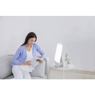 Eine Frau sitzt auf einem Sofa und liest ein Buch. Neben ihr steht eine Lampe auf einem kleinen Tisch. Die Umgebung ist hell und schlicht.